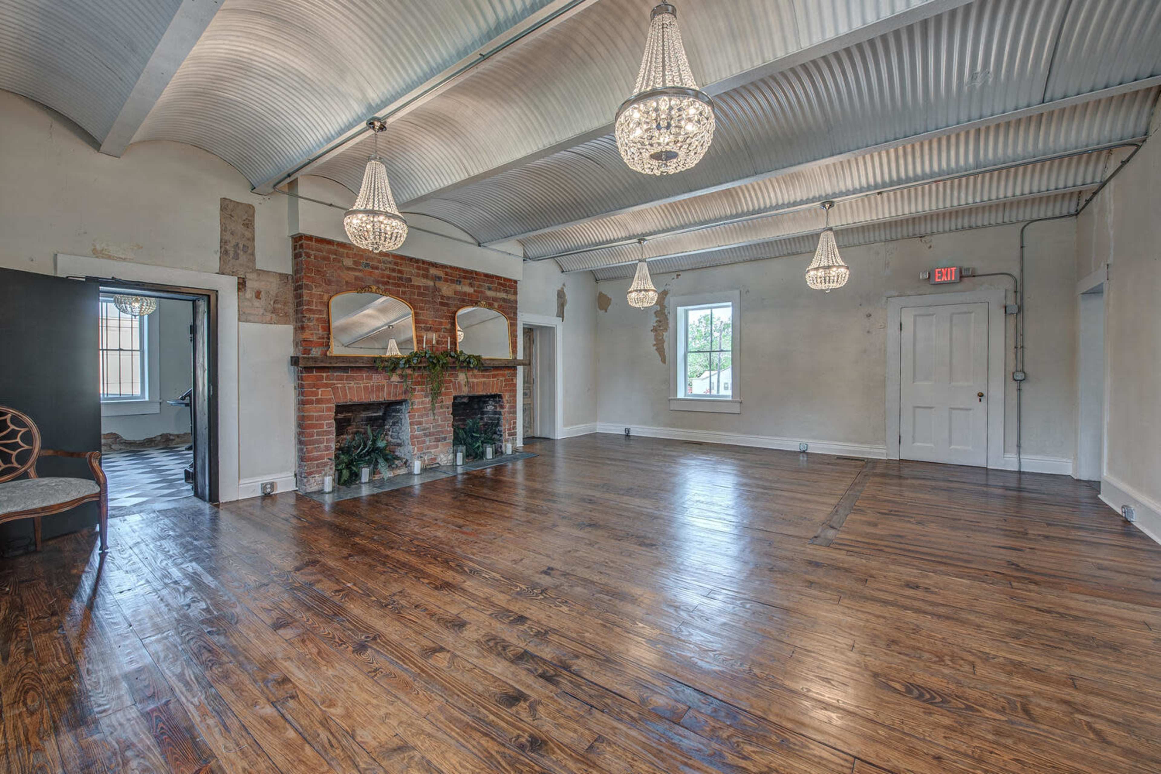 The image shows a spacious room with hardwood flooring, a brick fireplace, and three hanging chandeliers.