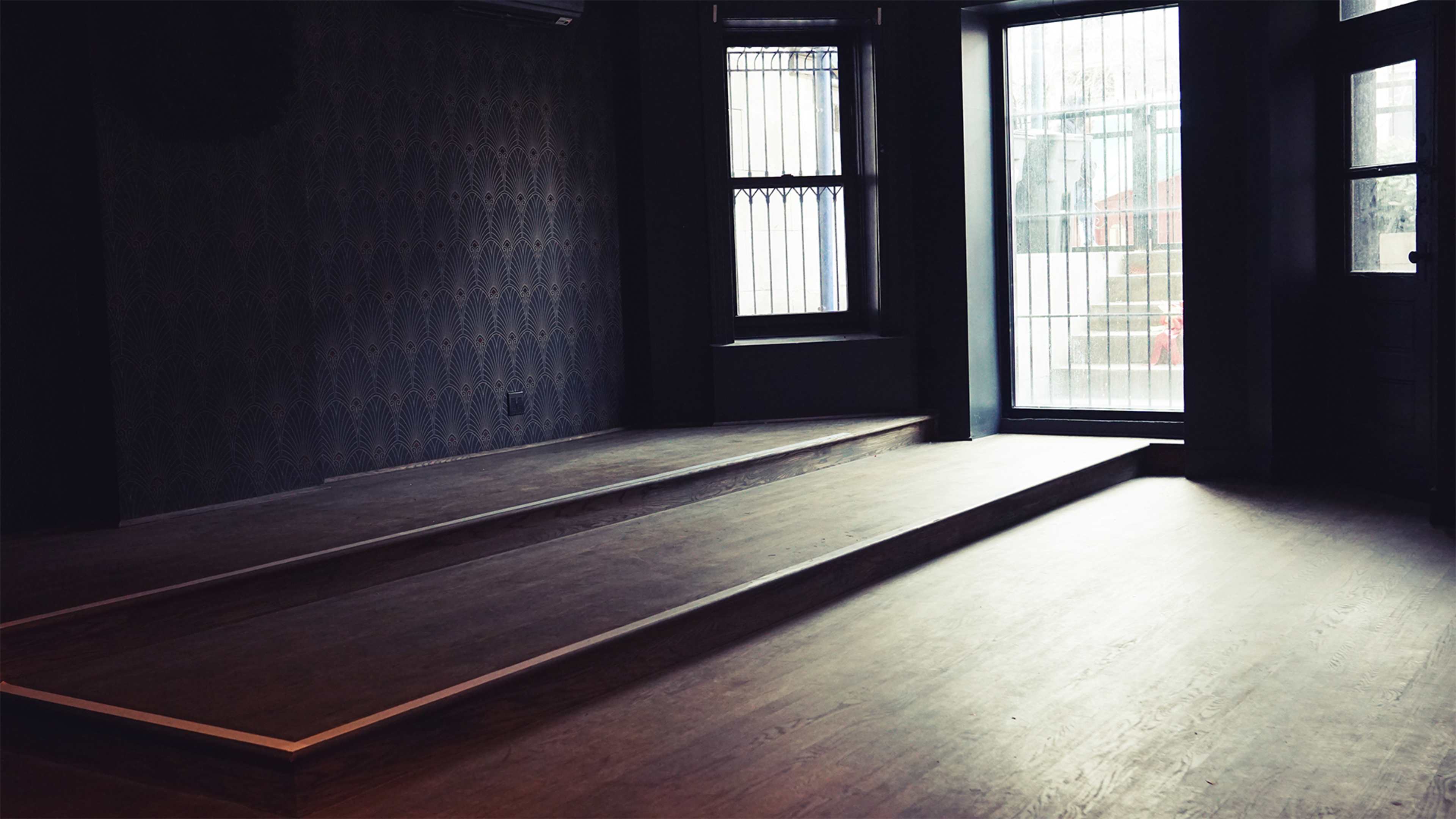 A dimly lit room with a raised wooden platform and large windows letting in natural light.