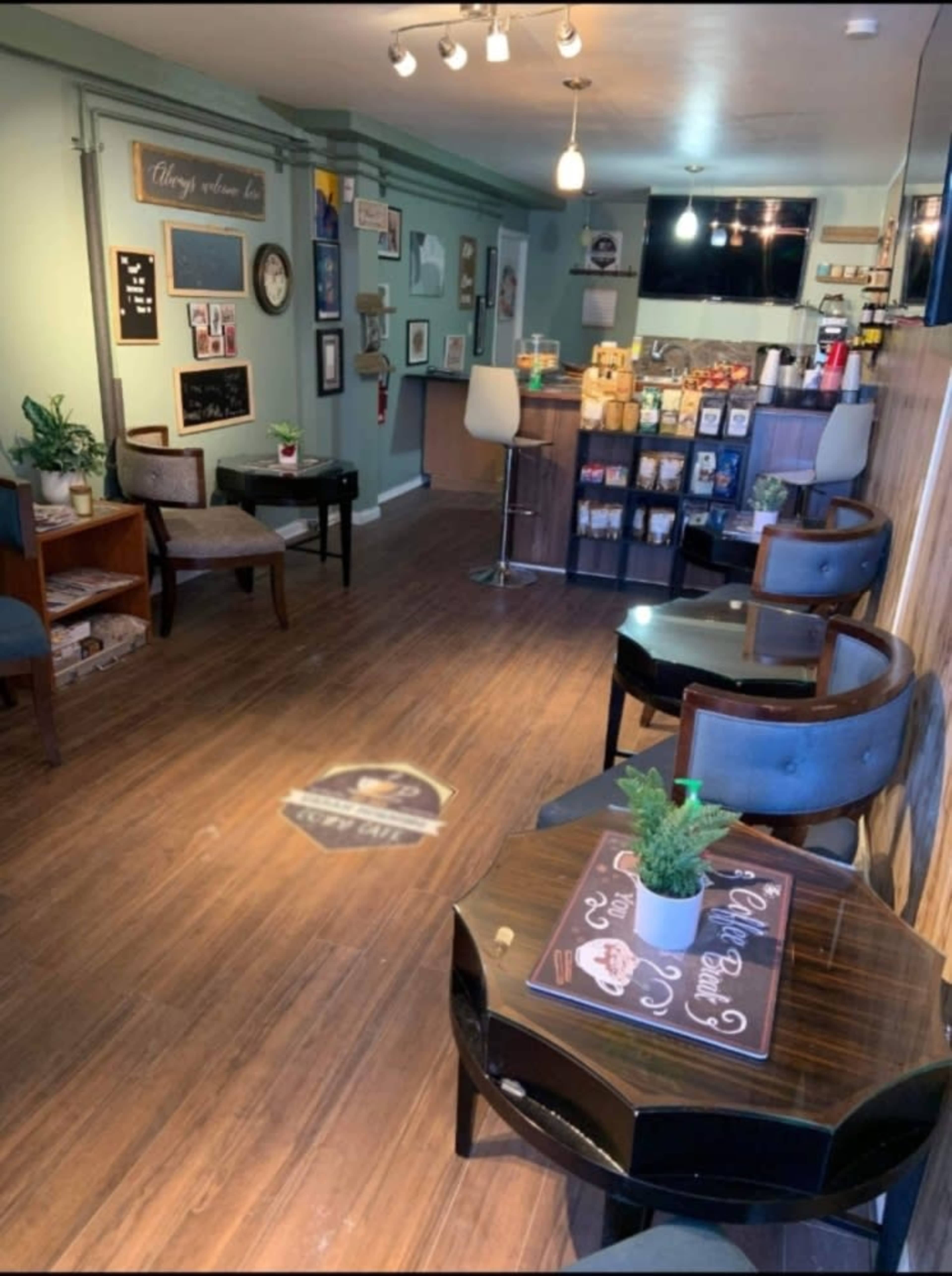 A cozy café interior with wooden flooring, a small seating area with chairs and tables, and a counter displaying various food and beverage items.