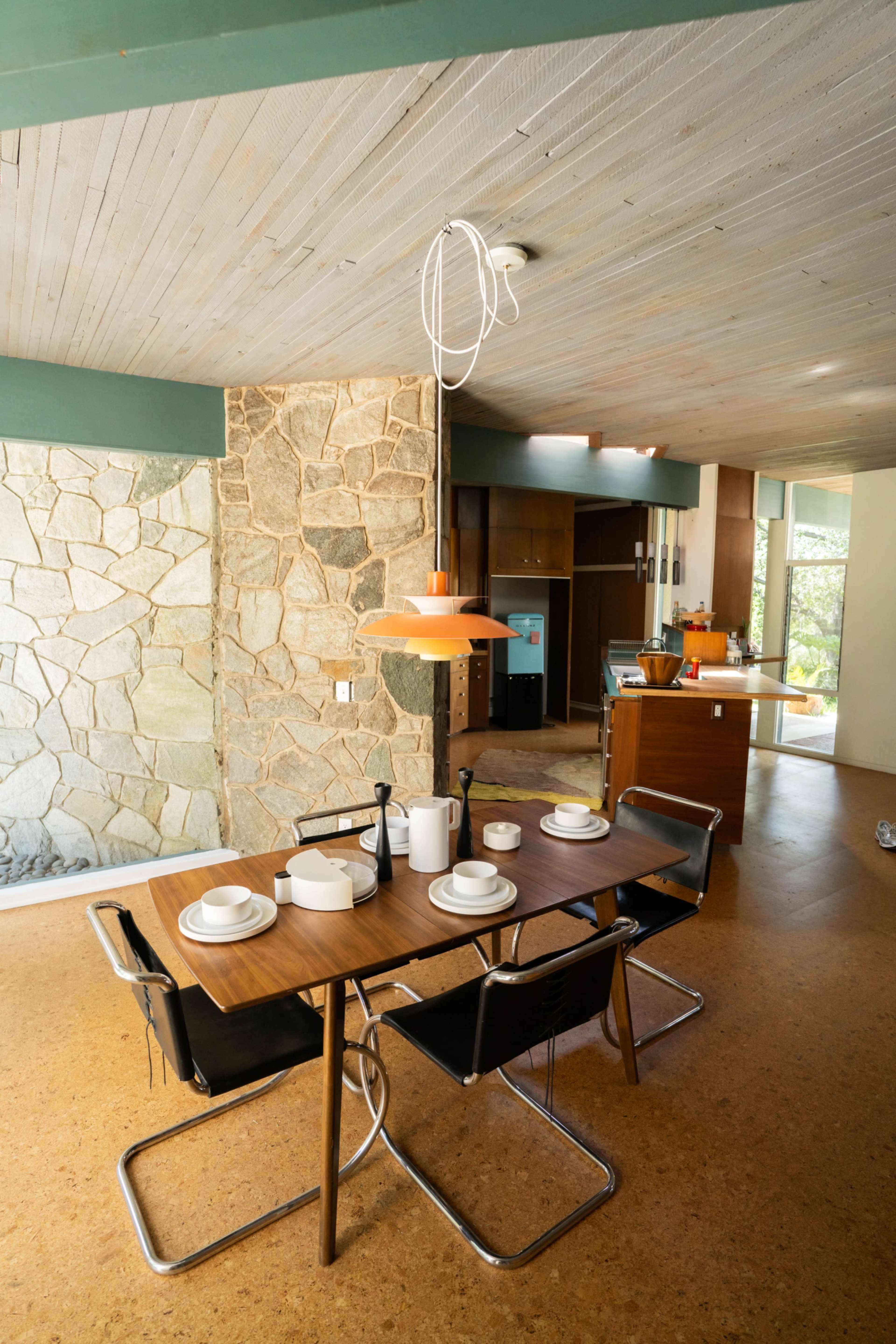 A dining table set with dishes and a pendant light hangs above, situated in a modern room featuring a stone wall and open kitchen.