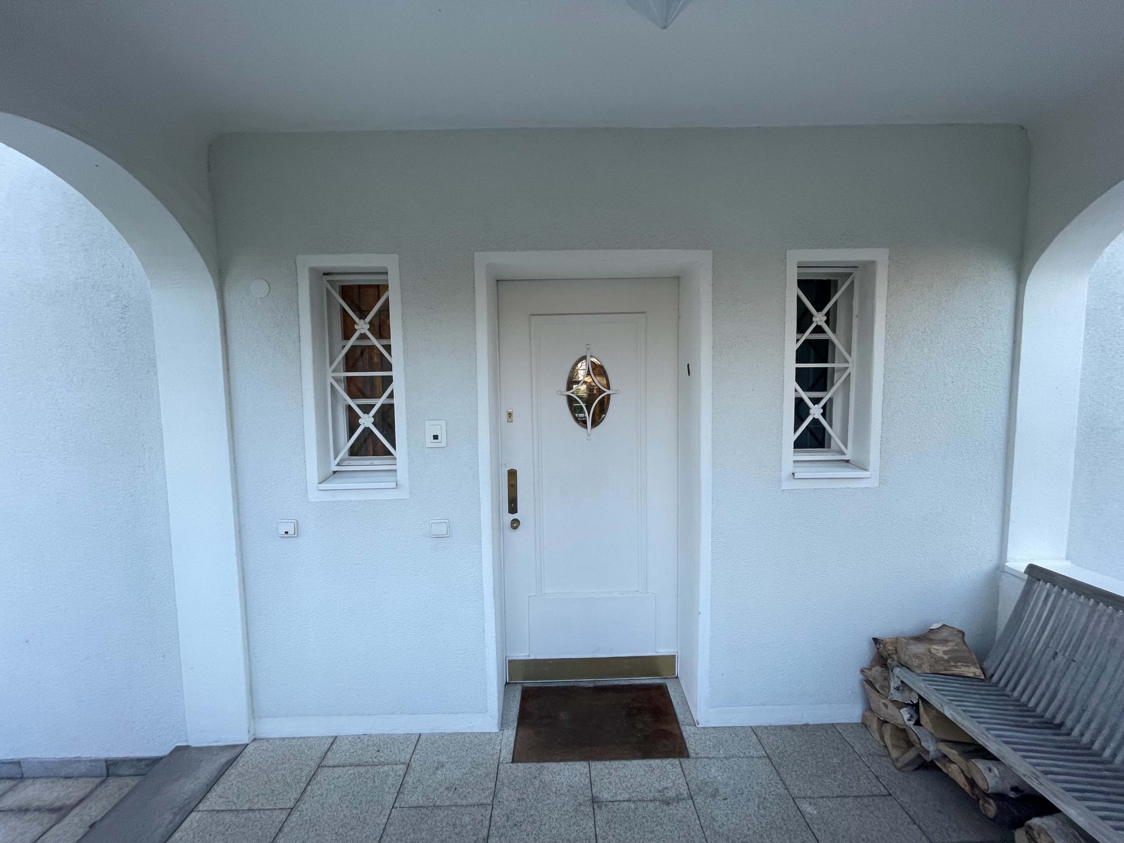A front entrance featuring a white door with a decorative window and two side windows, surrounded by a light-colored wall and a wooden bench with stacked firewood.
