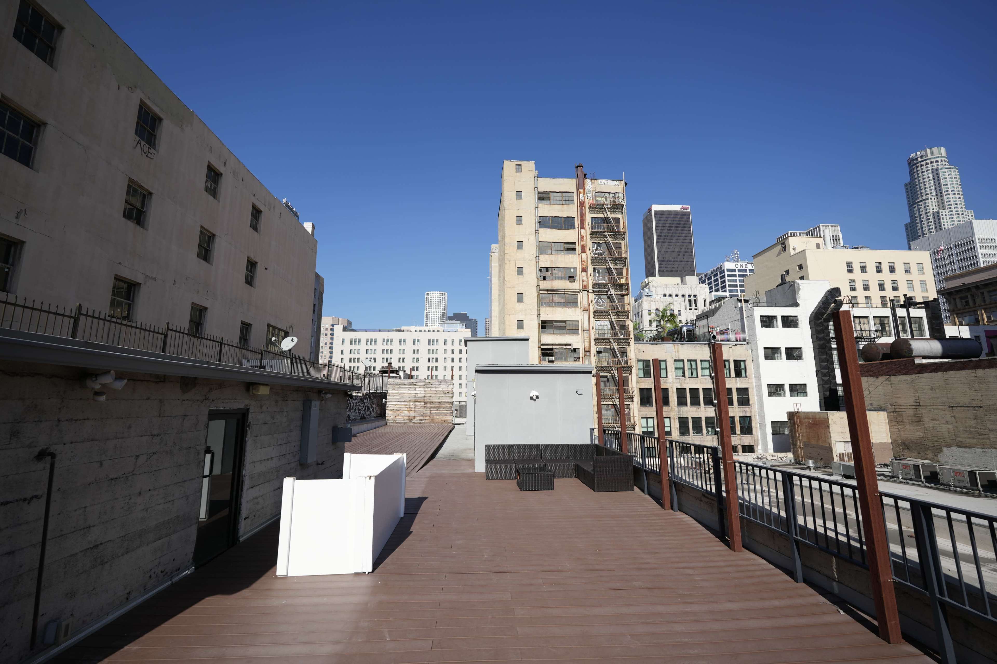 The image shows an urban rooftop with a wooden deck, featuring white furniture and a backdrop of high-rise buildings under a clear blue sky.