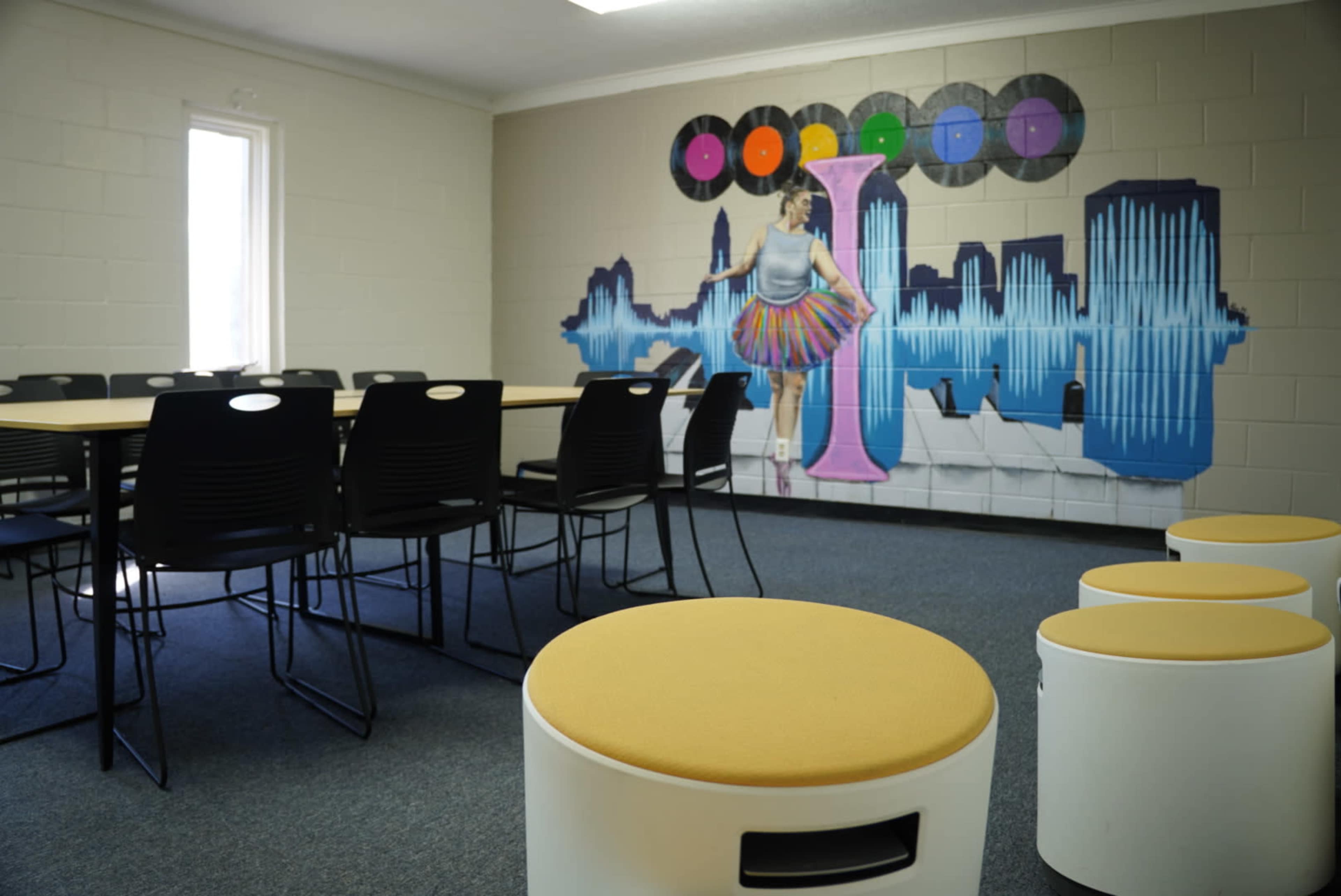 A classroom with a large table surrounded by black chairs and a mural of a woman in a colorful skirt against a soundwave backdrop on the wall.