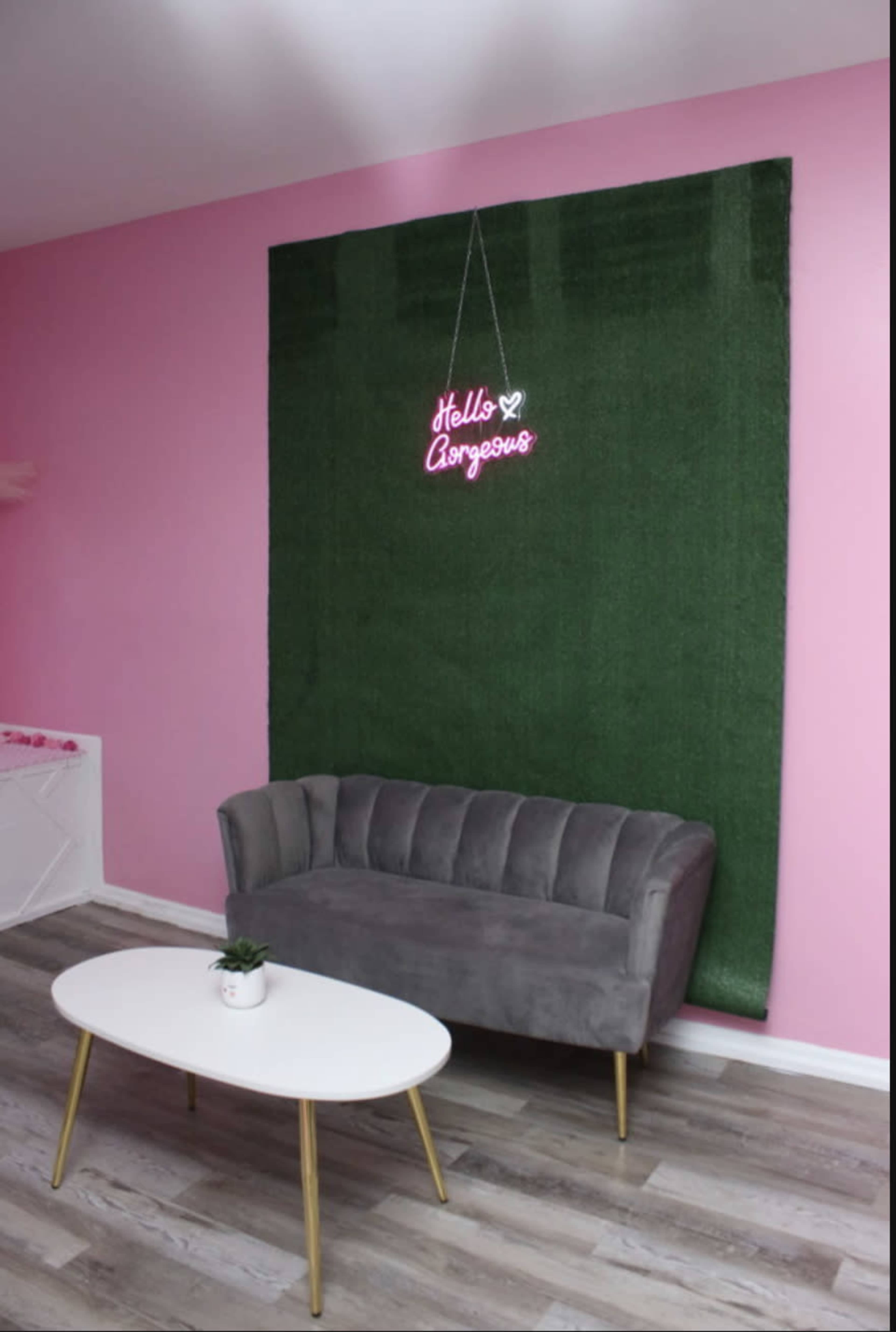 A gray couch placed in front of a green wall with a neon sign that says "Hello Gorgeous," against a pink backdrop.