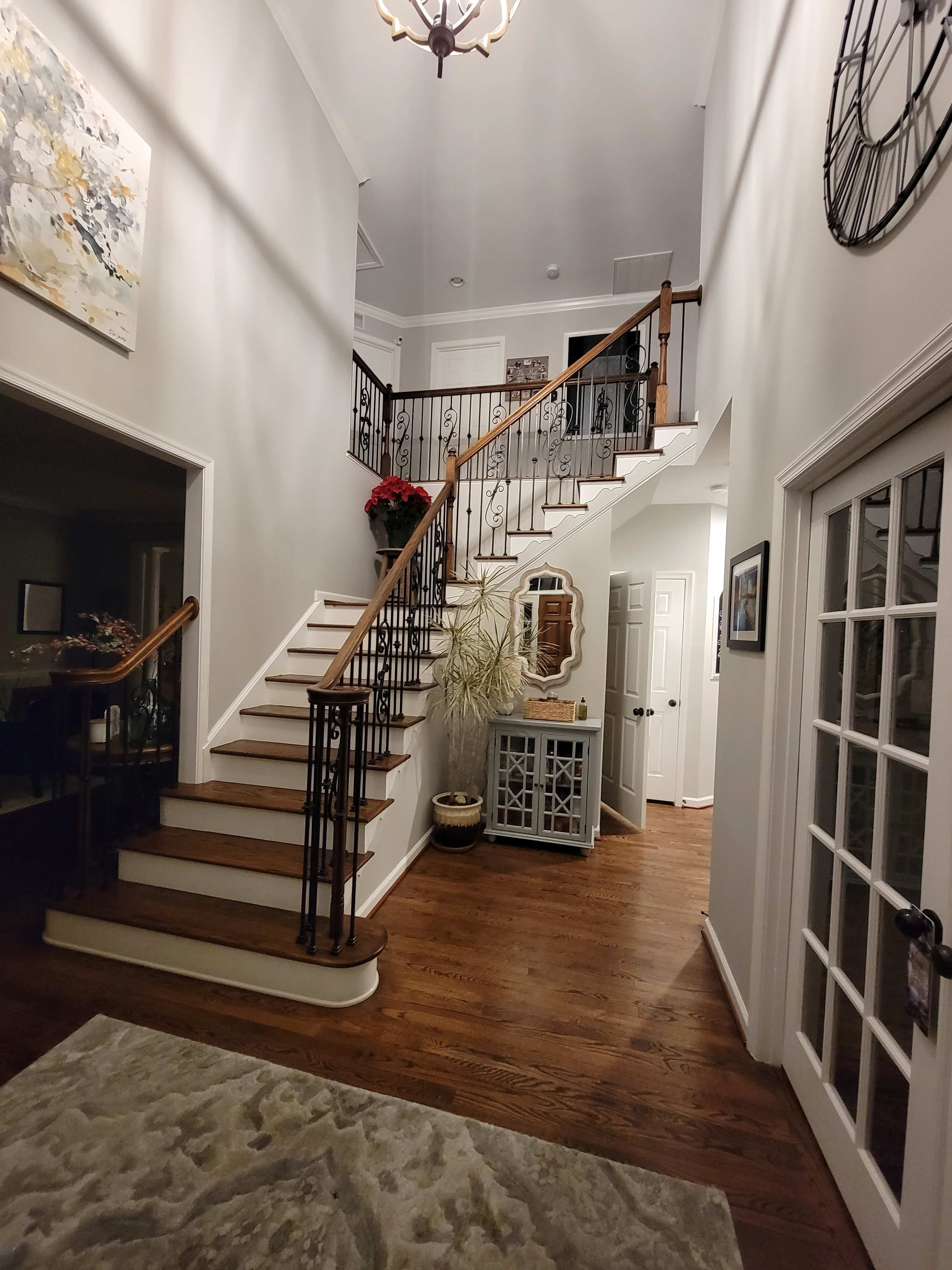A spacious foyer featuring a staircase with wrought-iron railings, flanked by a large mirror and a decorative plant, with warm wooden flooring throughout.