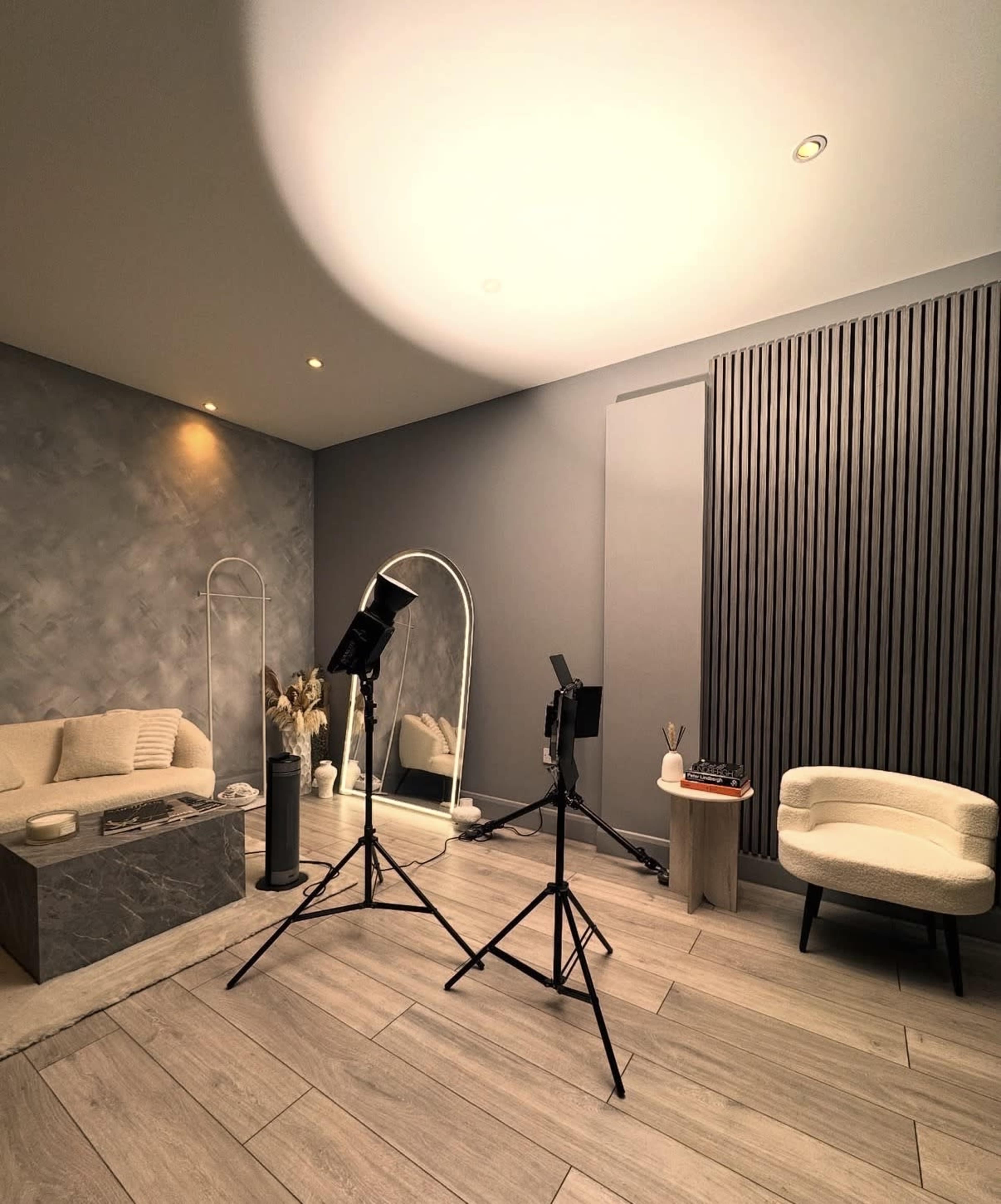 The image depicts a modern indoor space featuring a stylish seating area, a large mirror, and photography equipment set up on a hardwood floor.