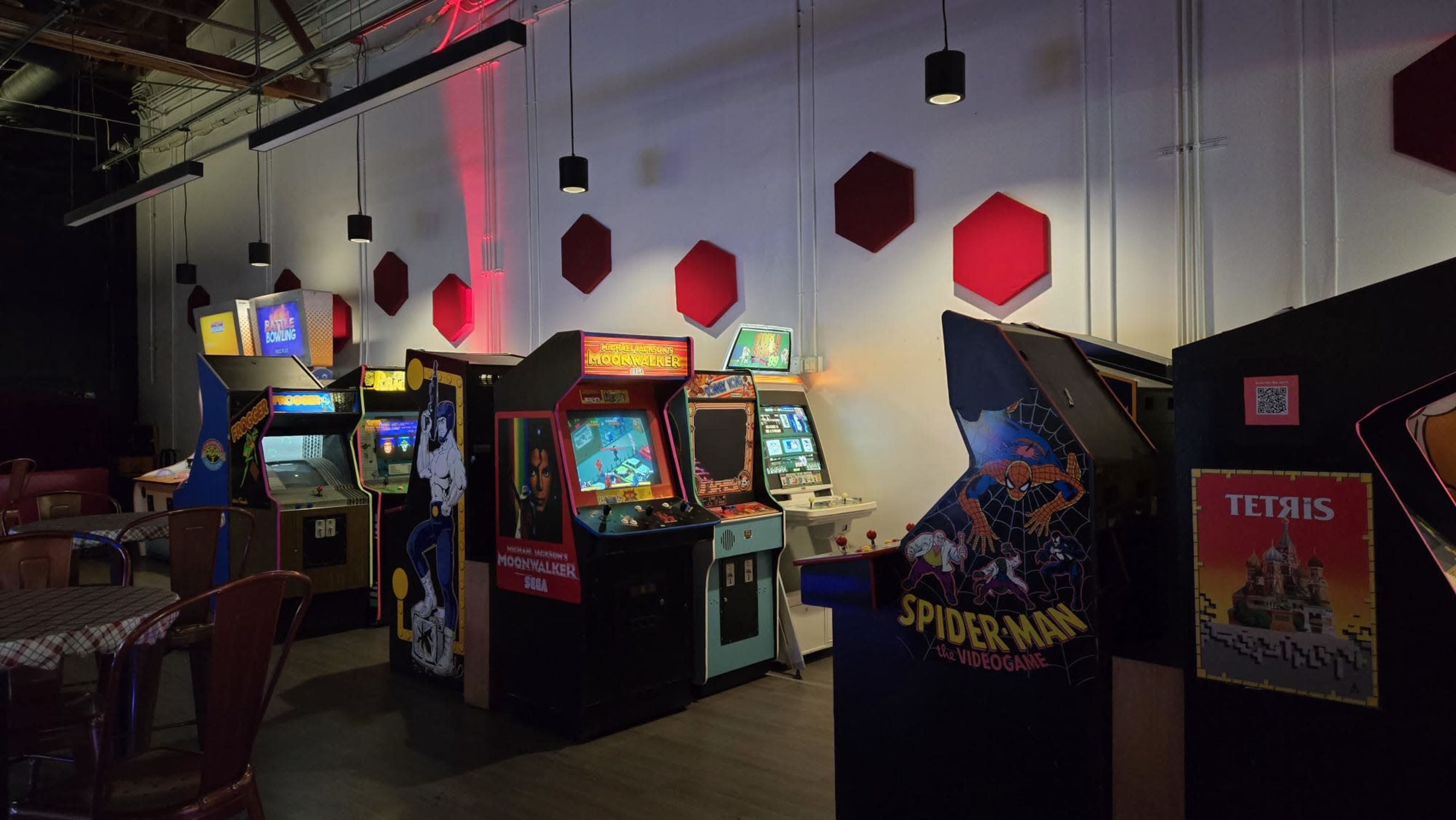 The image shows a row of vintage arcade machines displayed against a white wall with red hexagonal accents, illuminated in dim lighting.