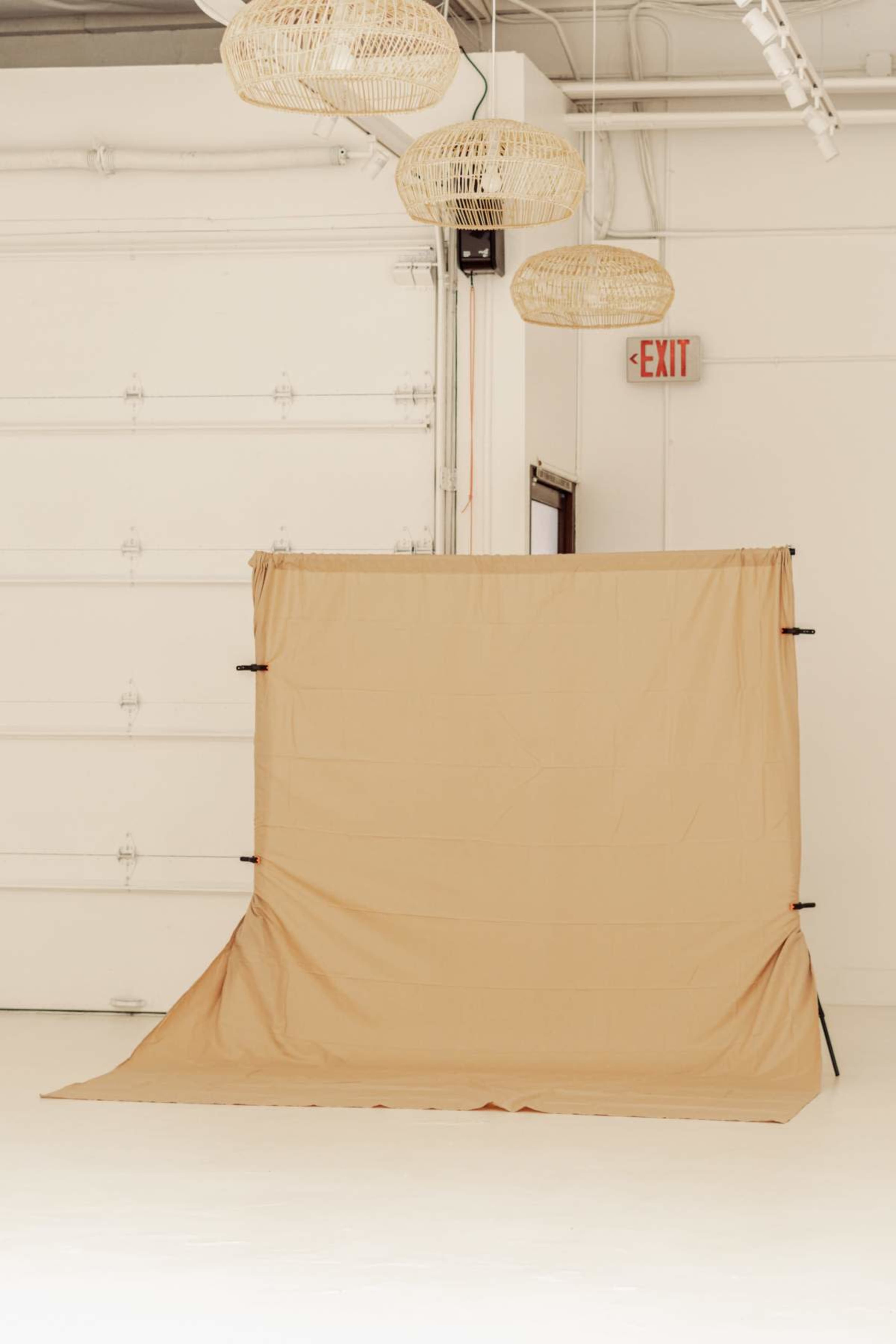 The image shows a plain beige backdrop on a stand in a well-lit studio space with woven light fixtures hanging from the ceiling.