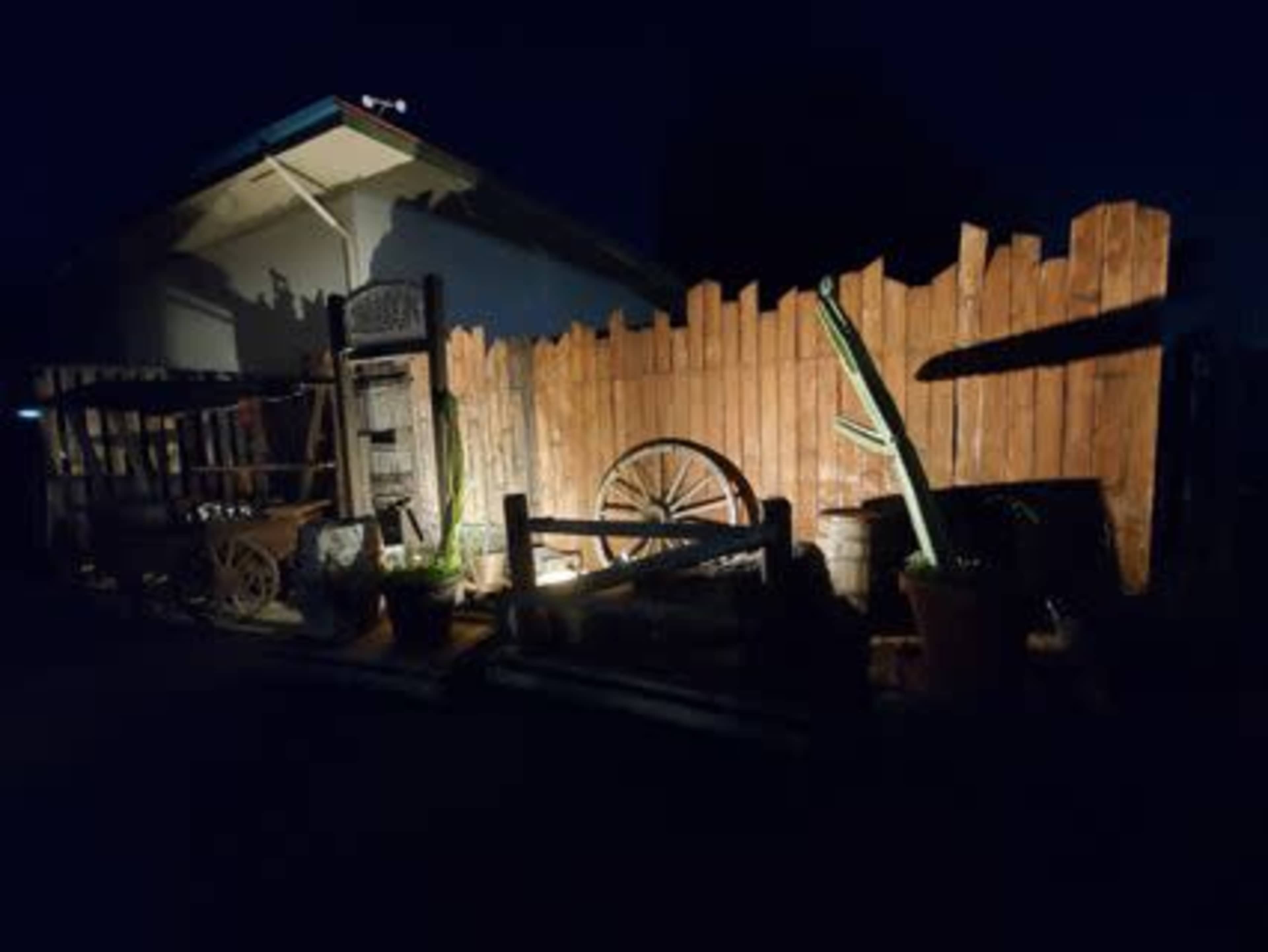 A dimly lit outdoor scene featuring wooden fences, a cartwheel, pots, and a cactus against a house at night.