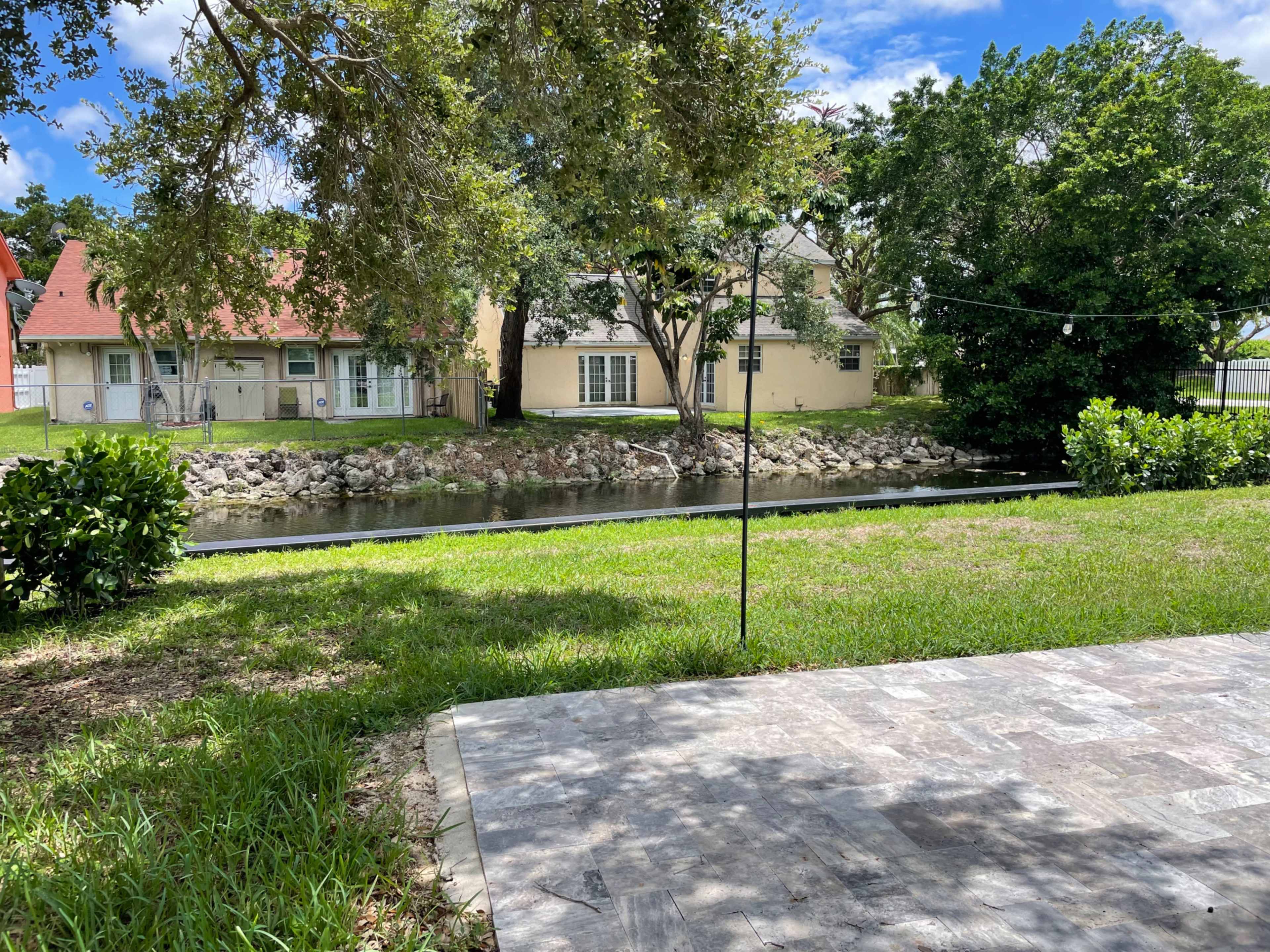 The image shows a grassy area with a stone patio overlooking a narrow waterway lined by trees and houses on both sides.