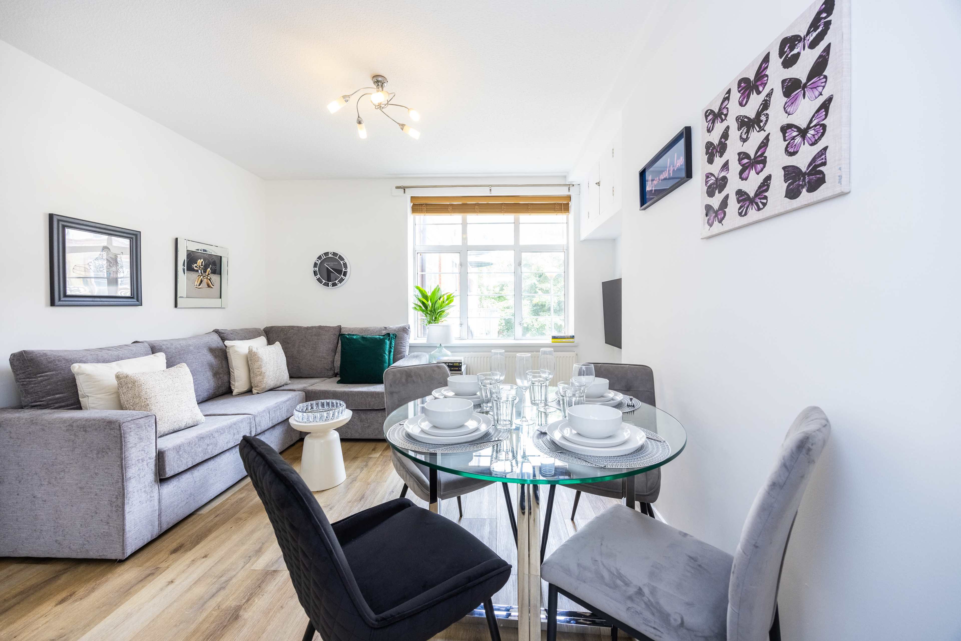 The image shows a modern living area with a gray sofa, a glass dining table set for four, and a window letting in natural light.