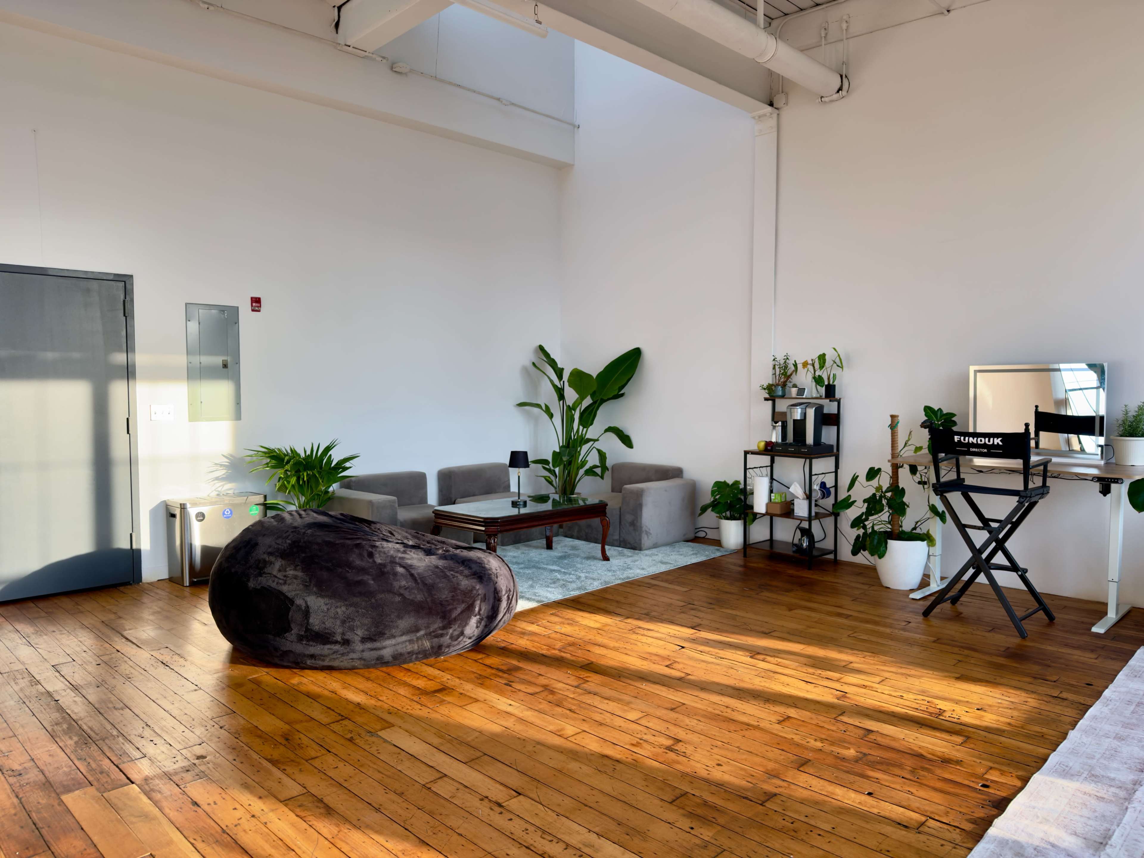 The image shows a spacious, well-lit room with wooden flooring, featuring a large bean bag chair, a seating area with a sofa and table, and some potted plants.