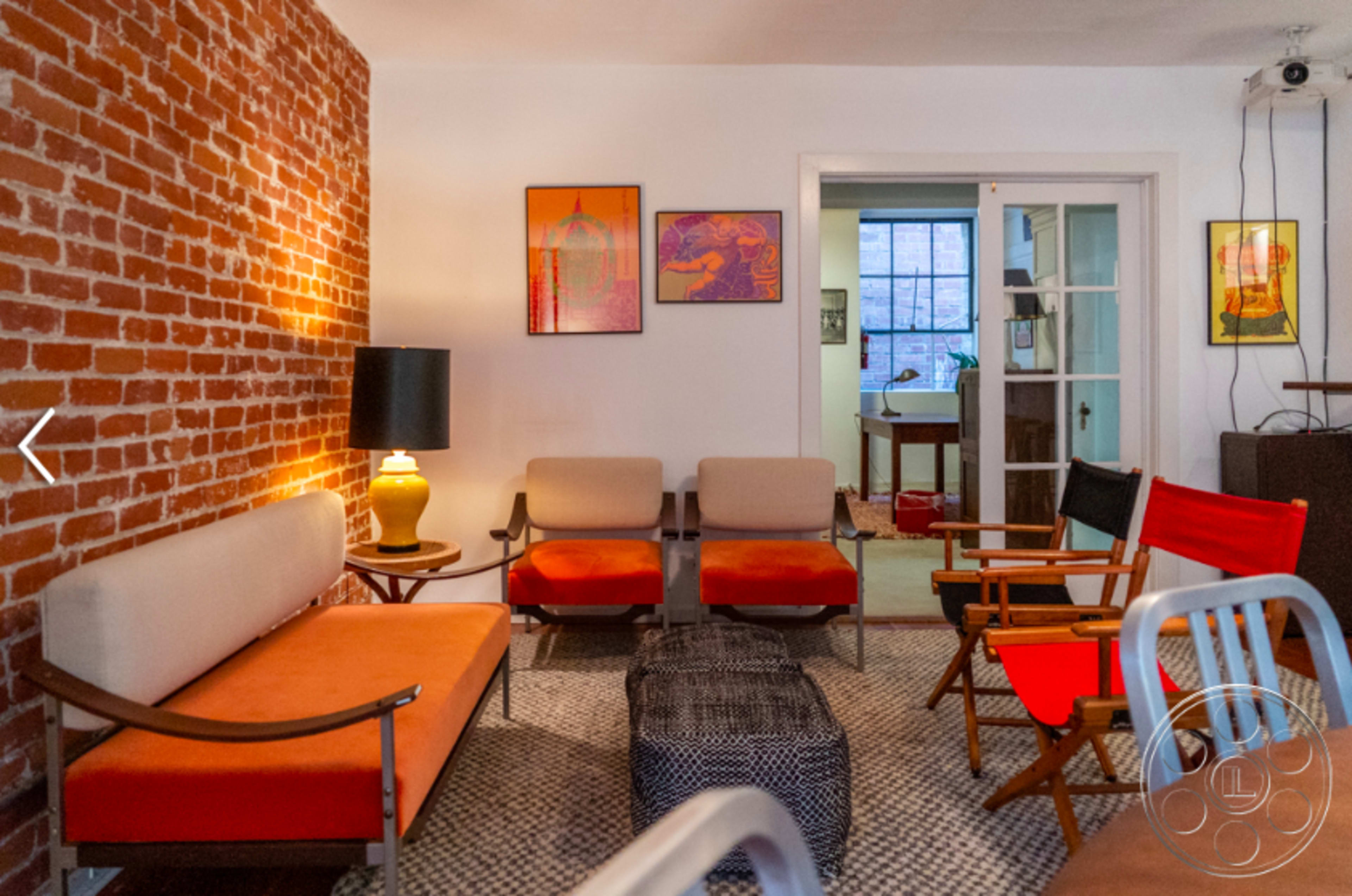 The image features a cozy living space with exposed brick walls, orange and gray furniture, and artwork on the walls.