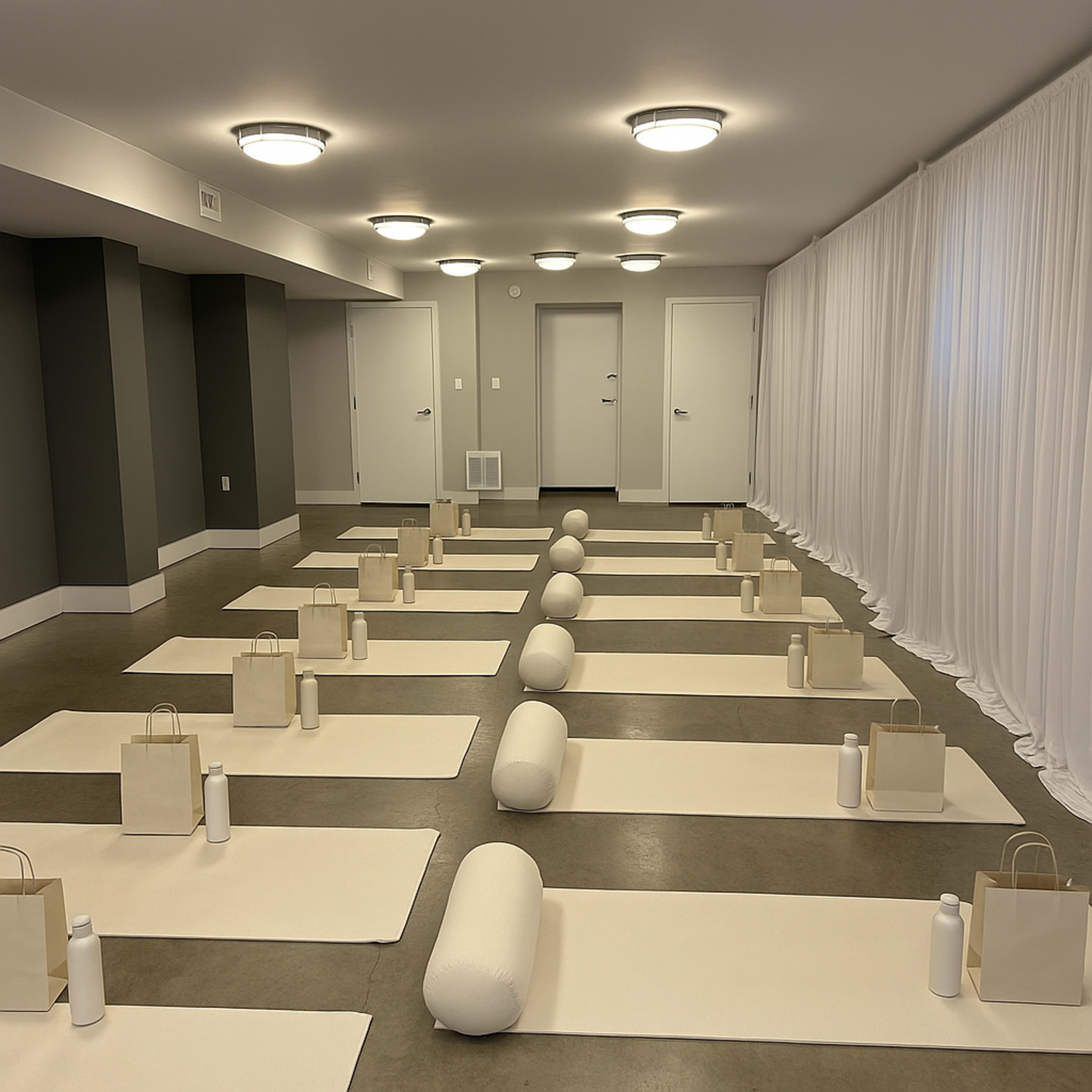 The image shows a yoga studio with neatly arranged yoga mats, bolsters, water bottles, and bags set up for a class.