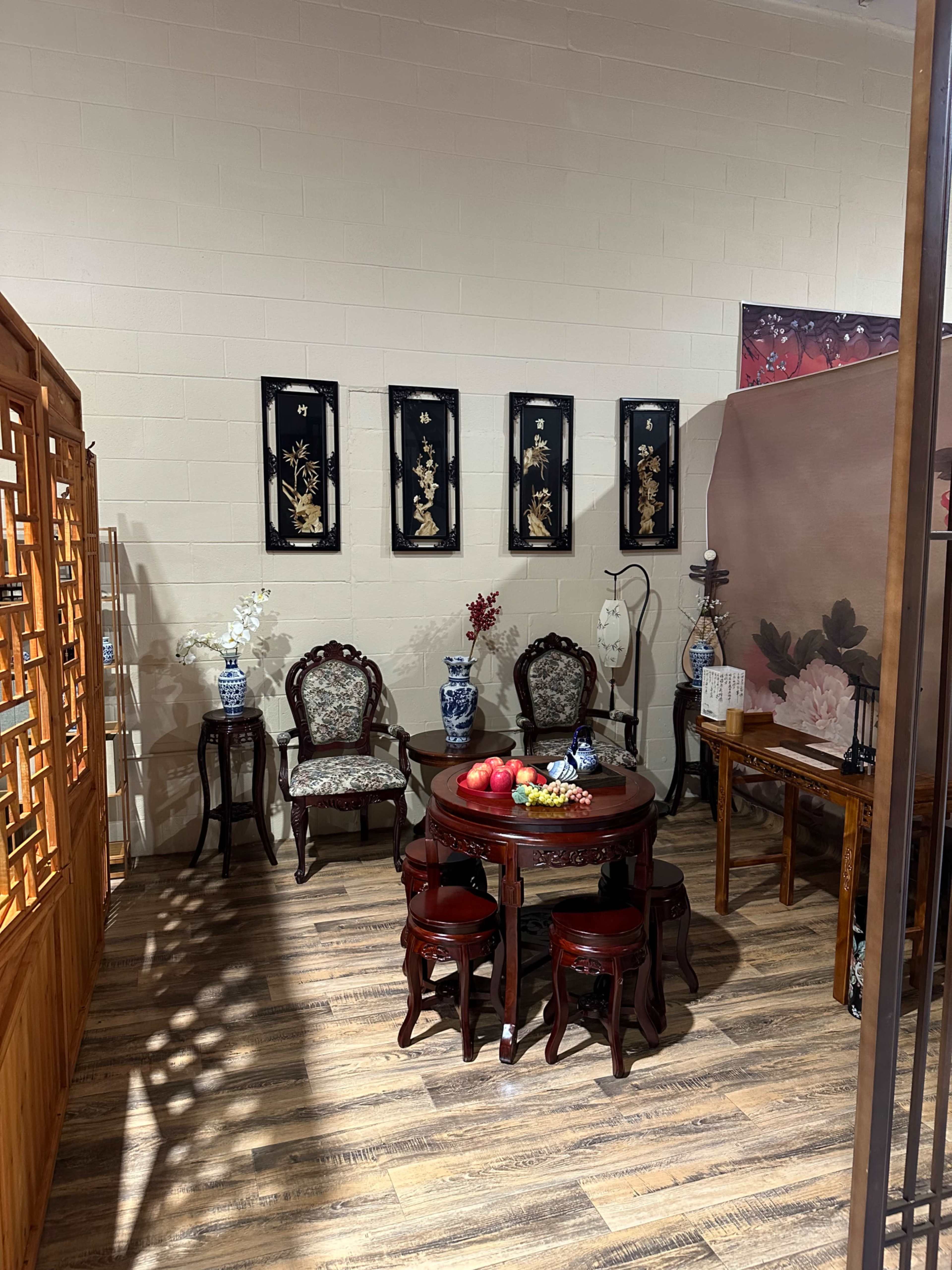 The image shows a traditional Asian-styled room featuring a round wooden table surrounded by ornate chairs, decorative vases, and framed artwork on the walls.