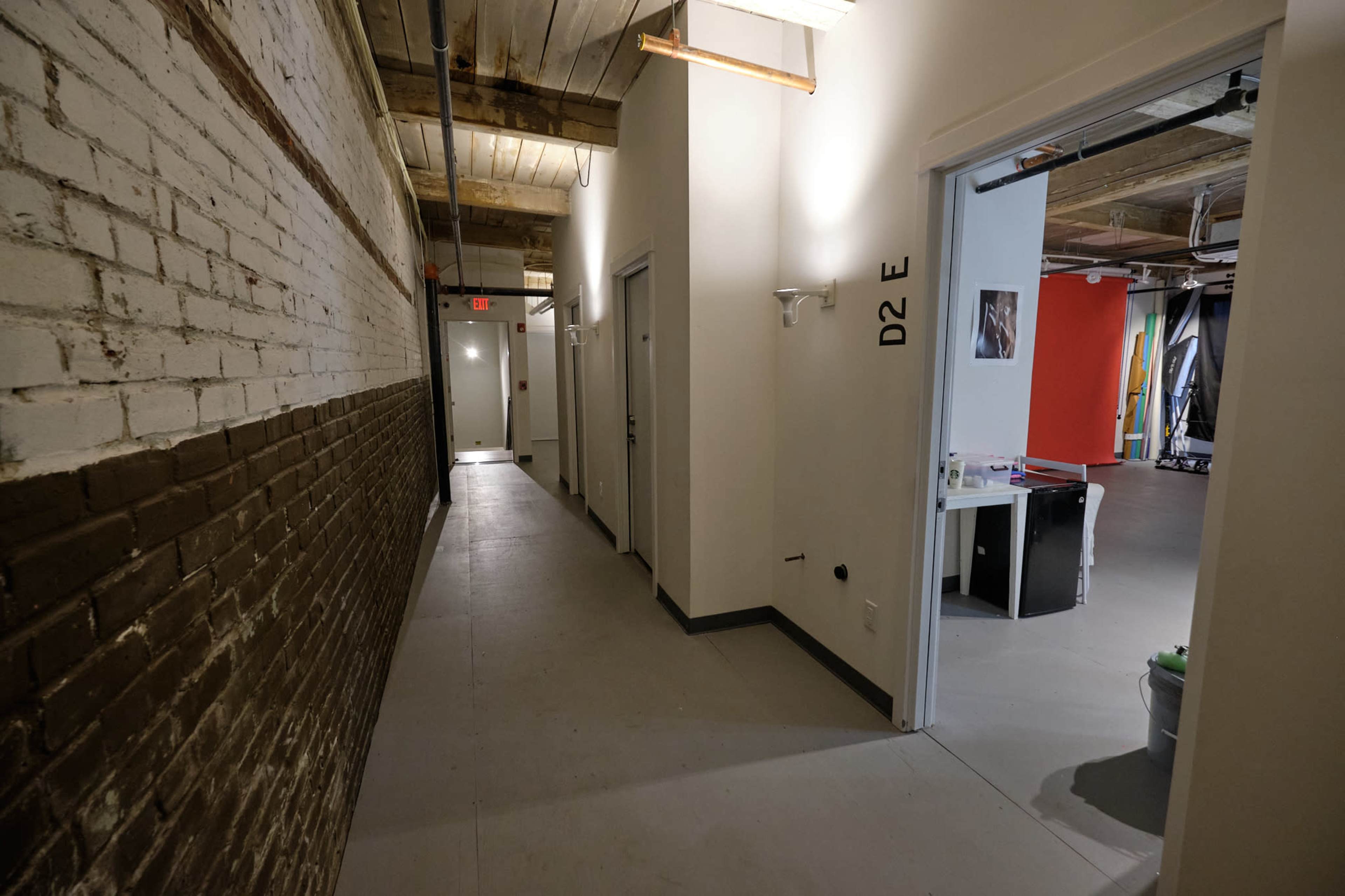 A long, narrow hallway with white walls and a dark brick section, leading to several doors on the left and an open workspace visible at the end on the right.