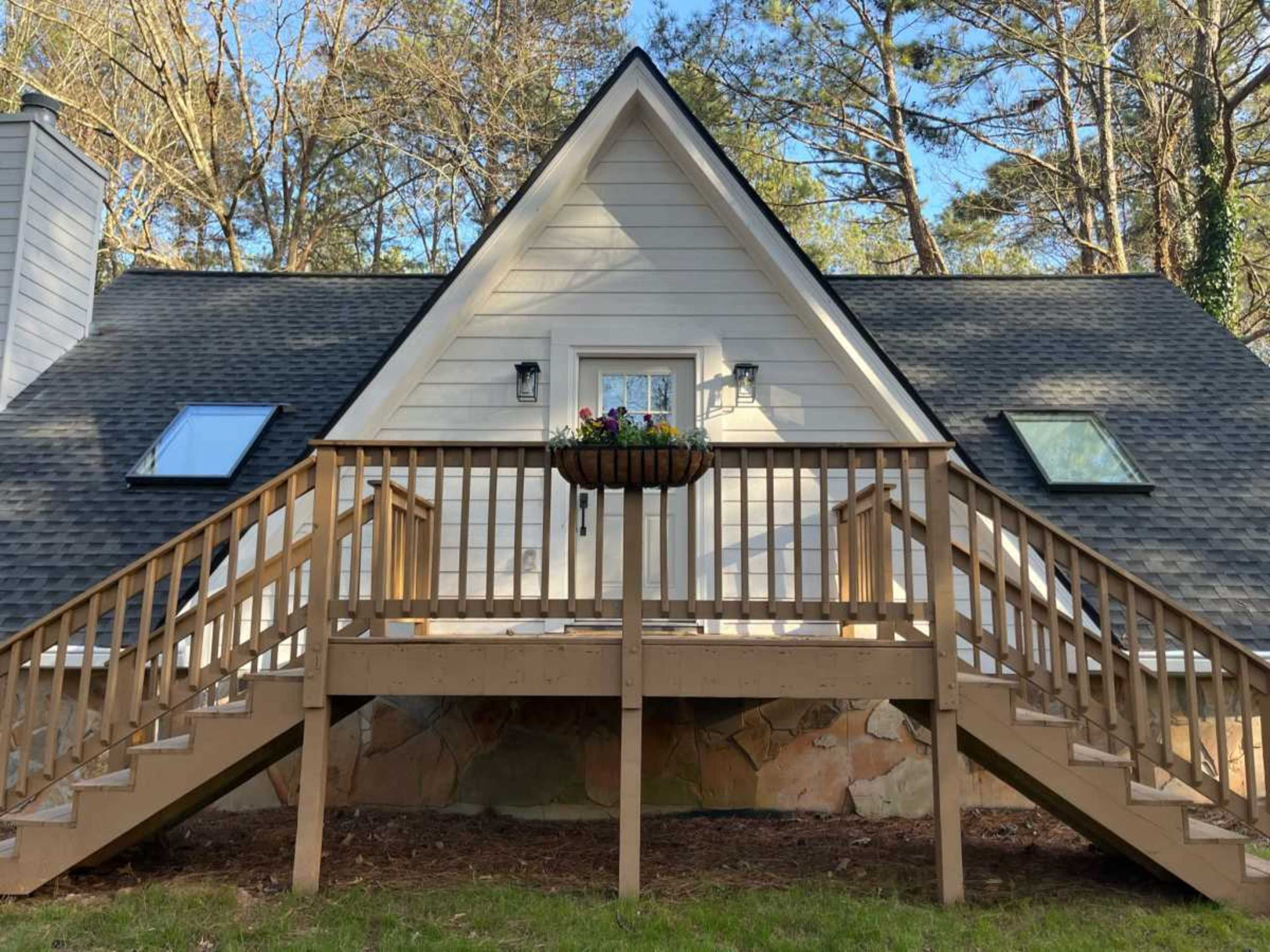 A triangular-roofed house with a wooden deck and stairs in a forested area.