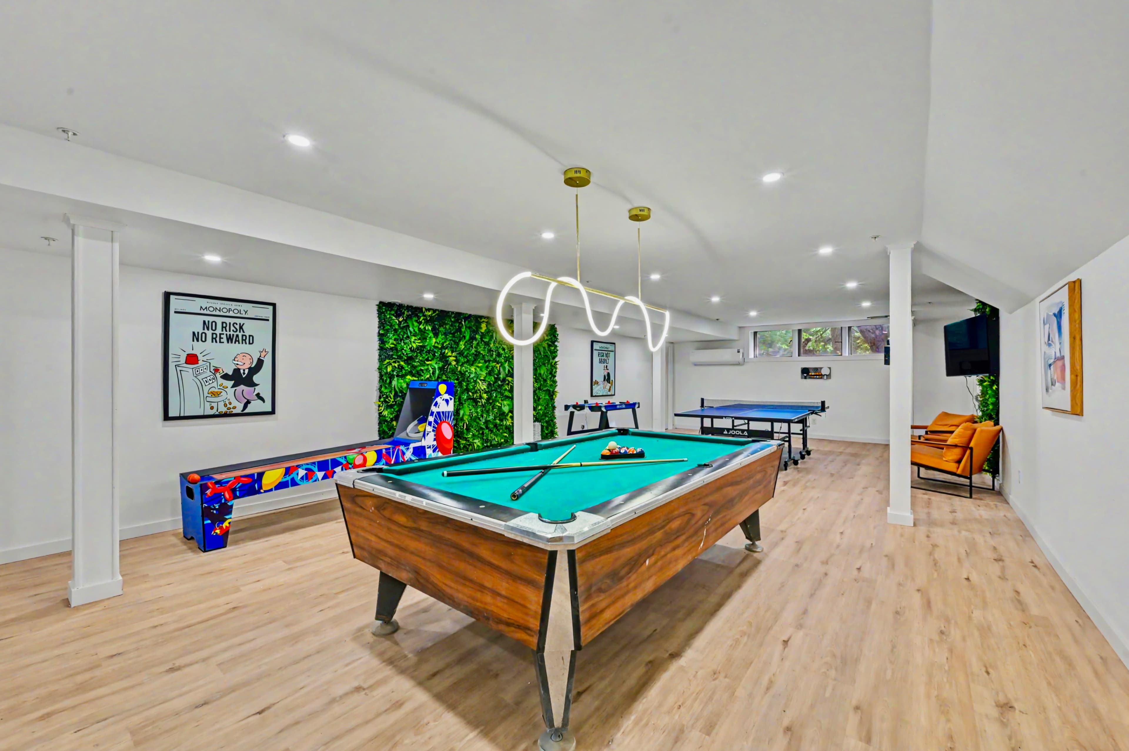 The image shows a recreational room featuring a pool table, arcade game, ping-pong table, and a seating area with an orange chair.