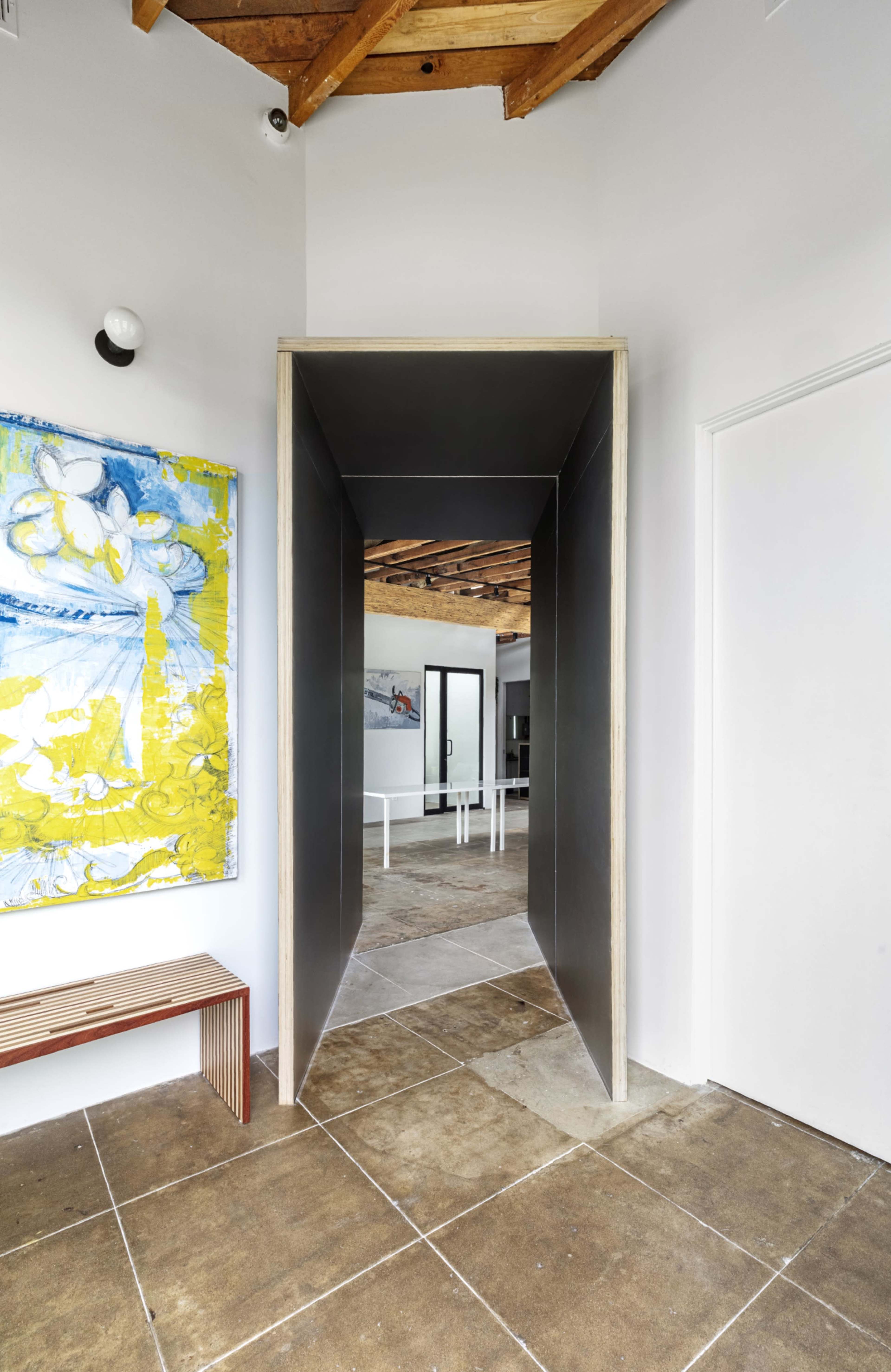 A hallway with a dark, framed entrance leads to a light-filled room featuring a wooden ceiling and a table set for dining.