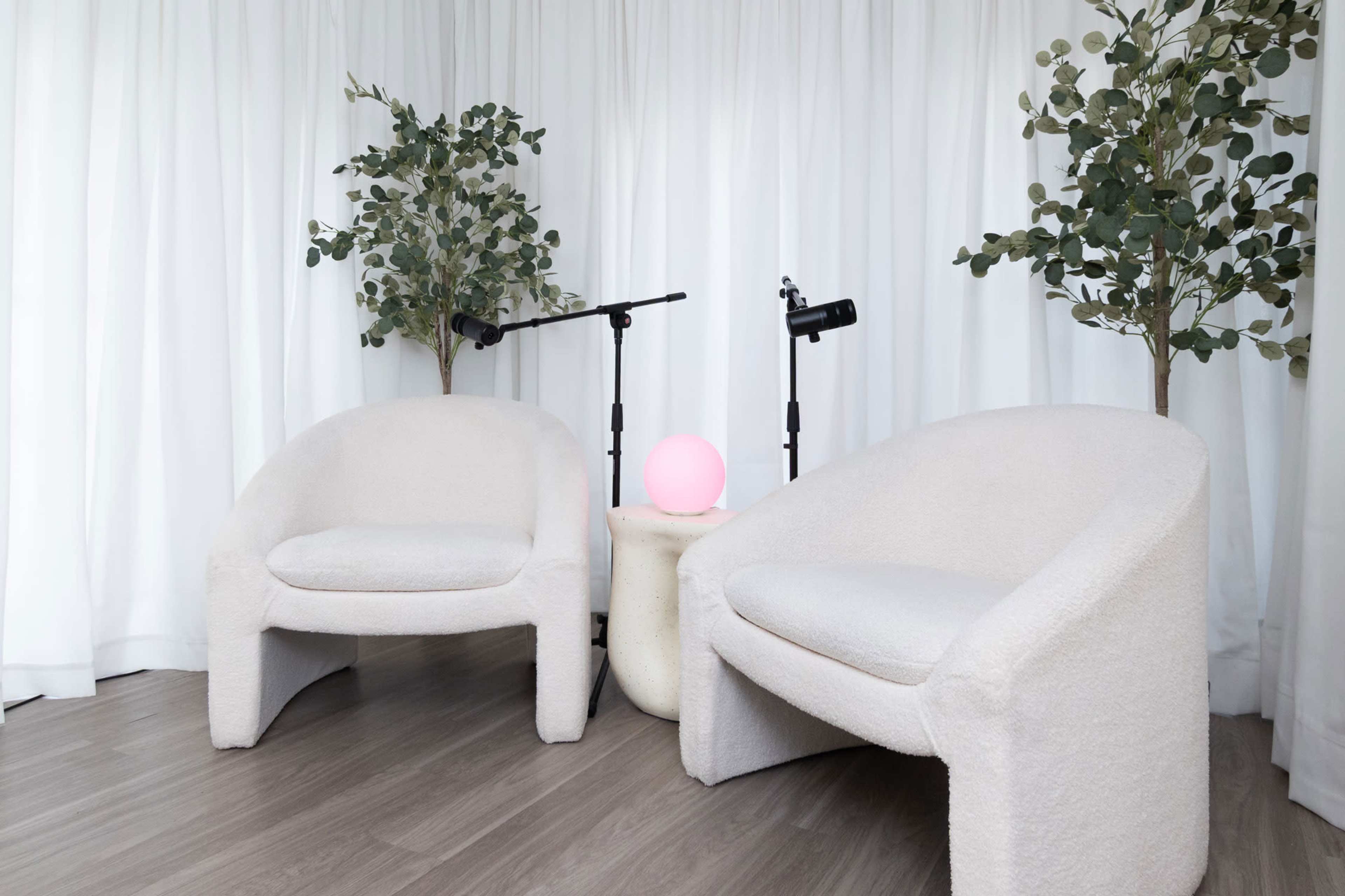 The image features two white, textured chairs arranged beside a small round table with a pink lamp, flanked by two potted plants, against a backdrop of white curtains.