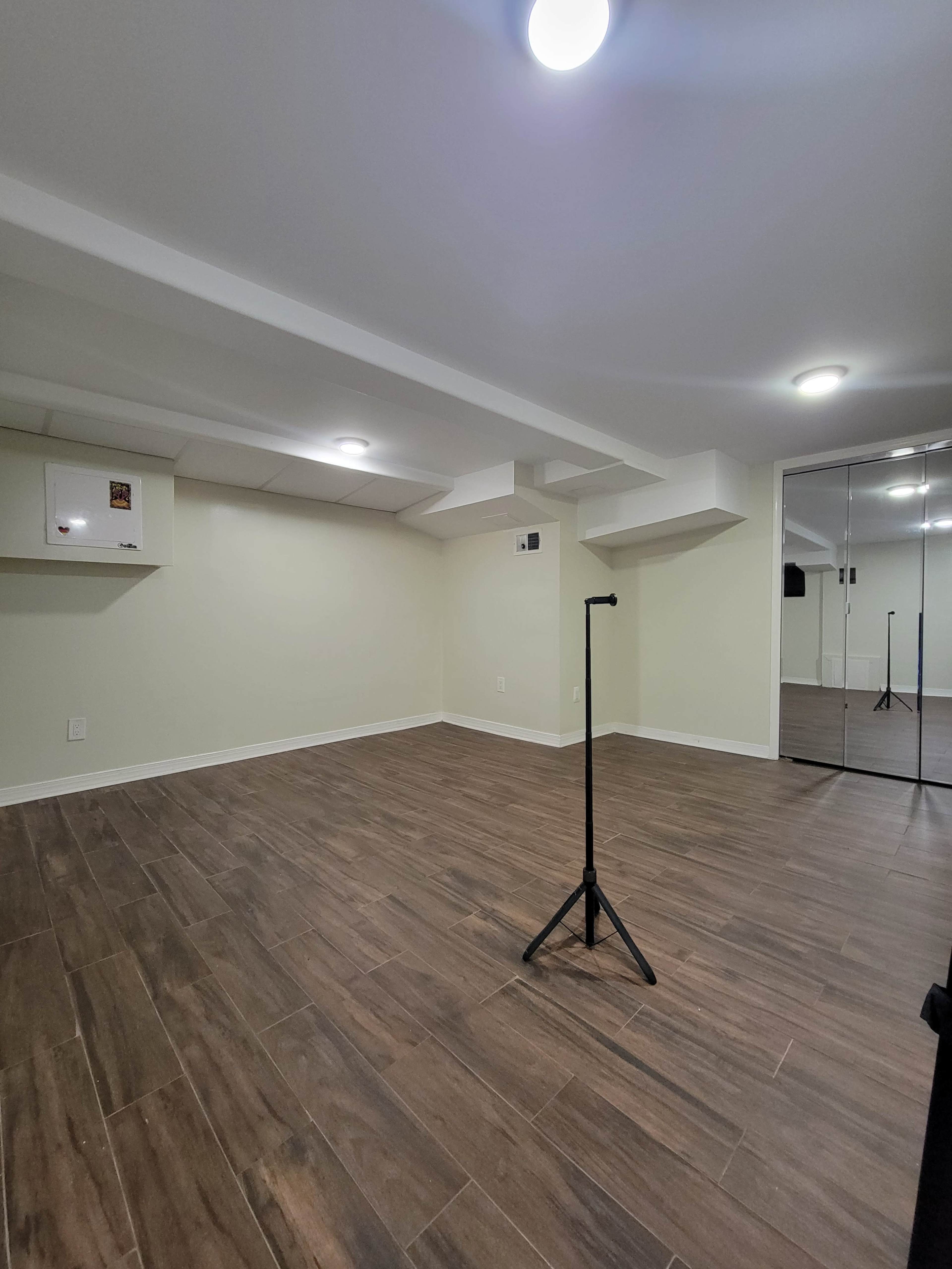 The image shows a spacious, well-lit basement room with wooden flooring and a mirrored wall.