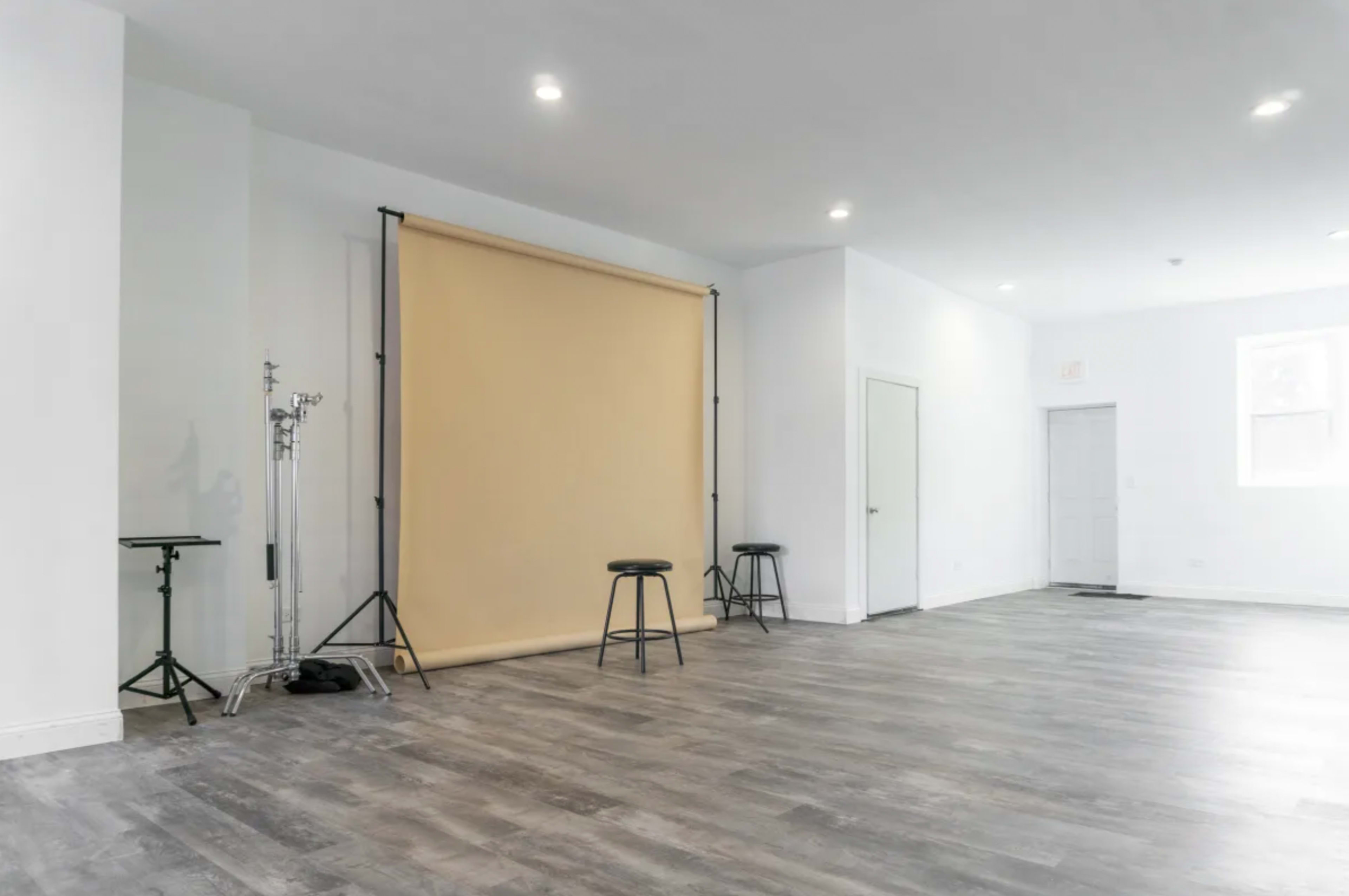 A spacious room with a beige backdrop set up on a stand, two black stools, and light-colored wooden flooring.