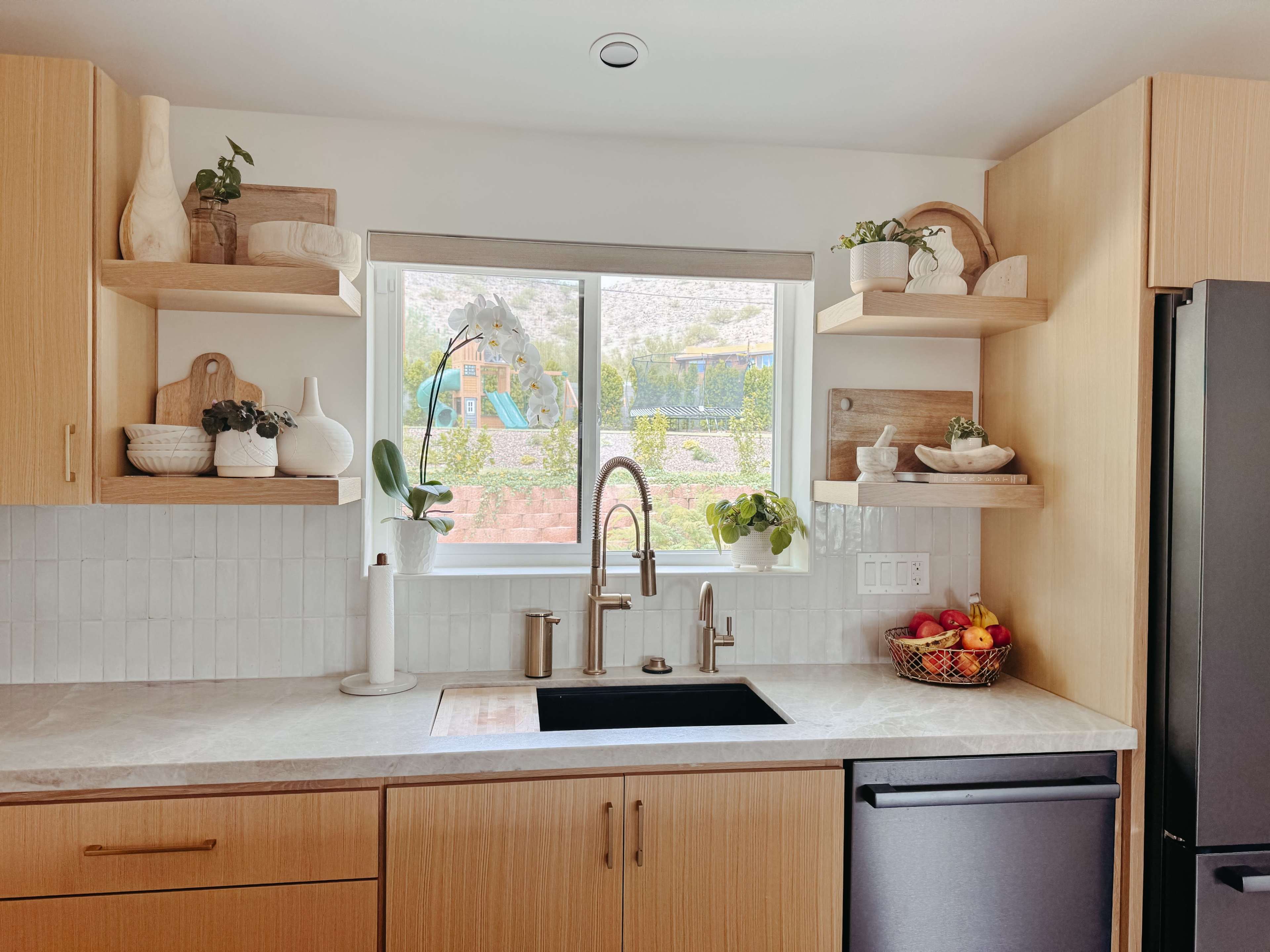 The image shows a modern kitchen with light wood cabinetry, a black sink, open shelves displaying decorative items and plants, and a window providing natural light.