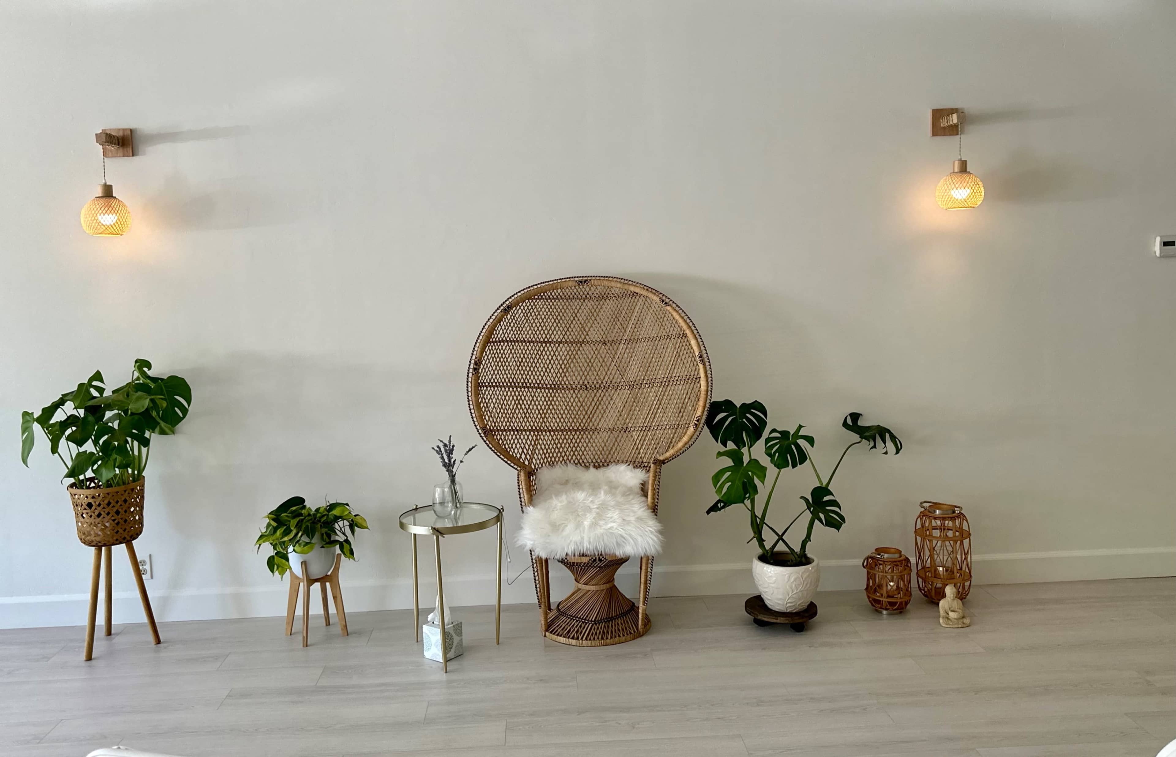 A minimalist interior featuring a rattan chair with a fur throw, flanked by various potted plants and decorative lighting on the wall.