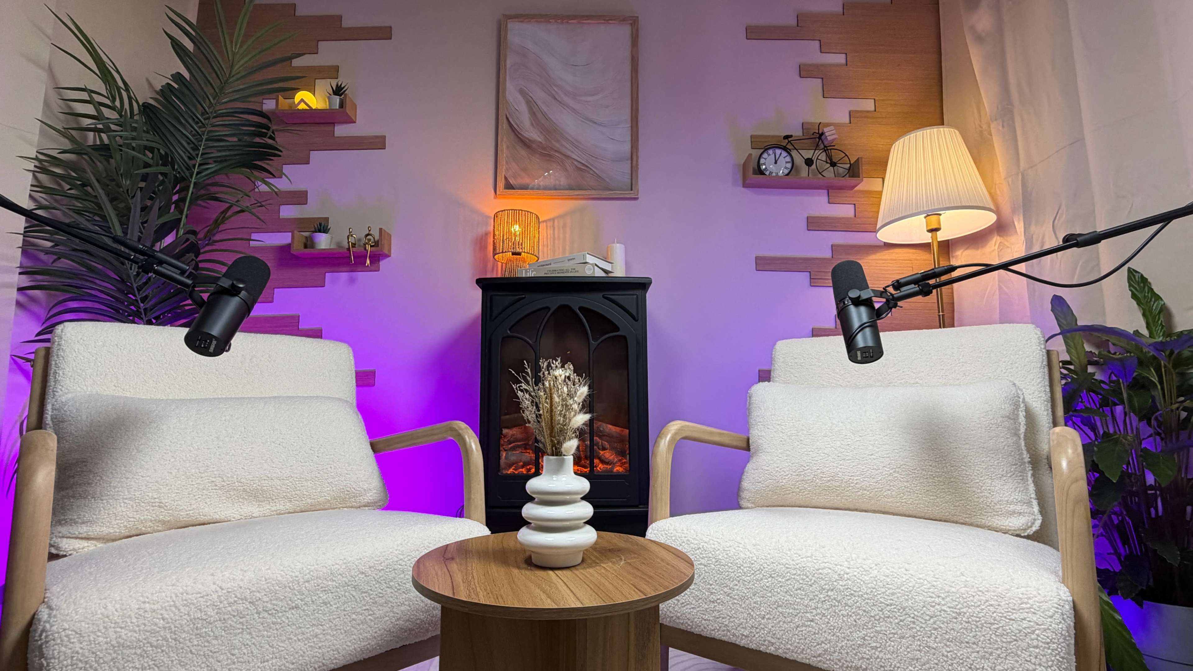 The image shows a cozy interior setup with two armchairs facing a small table, a decorative vase on the table, and a faux fireplace in the background illuminated by a warm light.