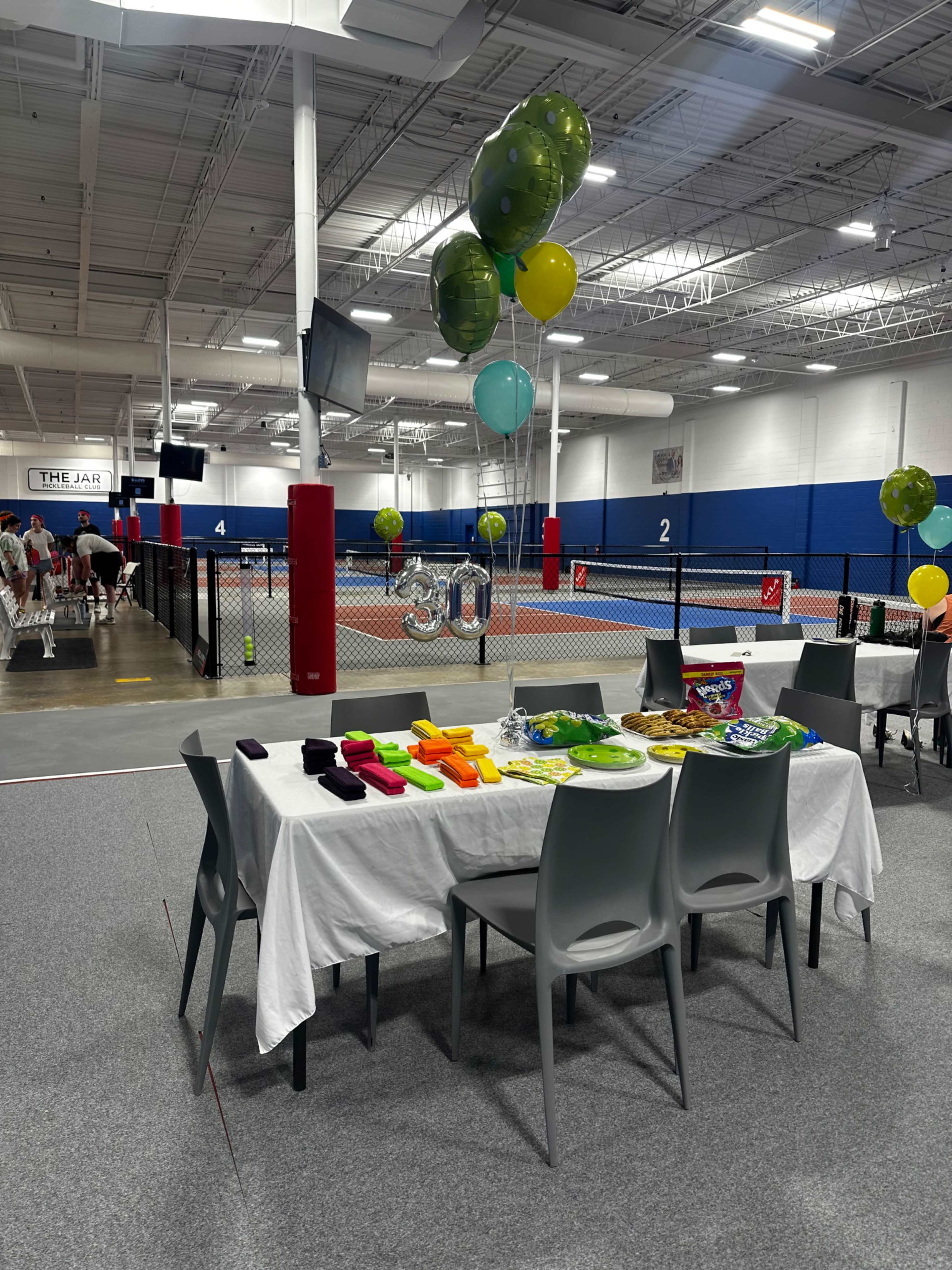 A tablescape with a white tablecloth is set with snacks and colorful balloons in a sports facility featuring tennis courts in the background.
