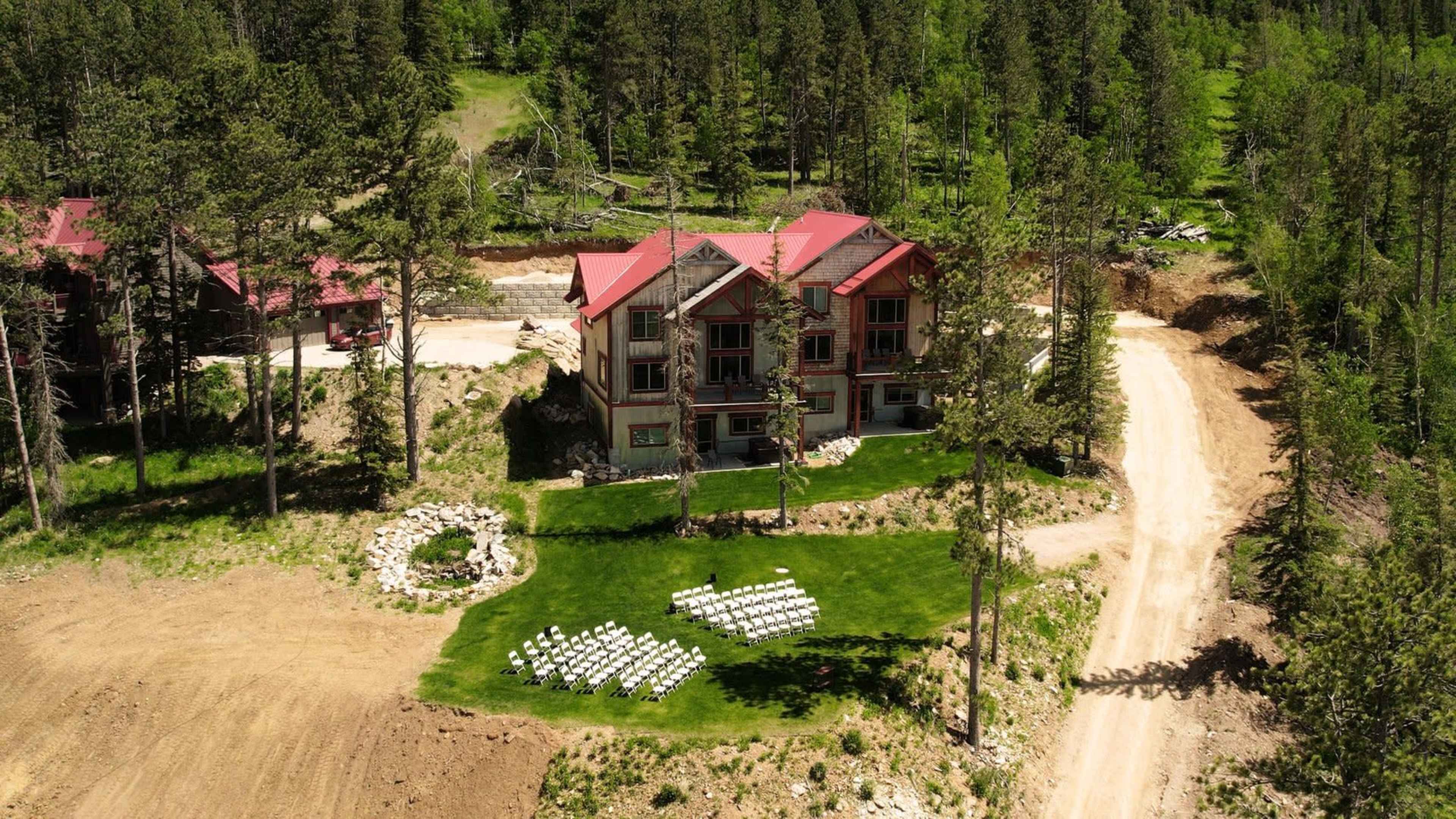 A large house with a red roof is surrounded by forest and a neatly arranged seating area on a green lawn, set for an outdoor event.