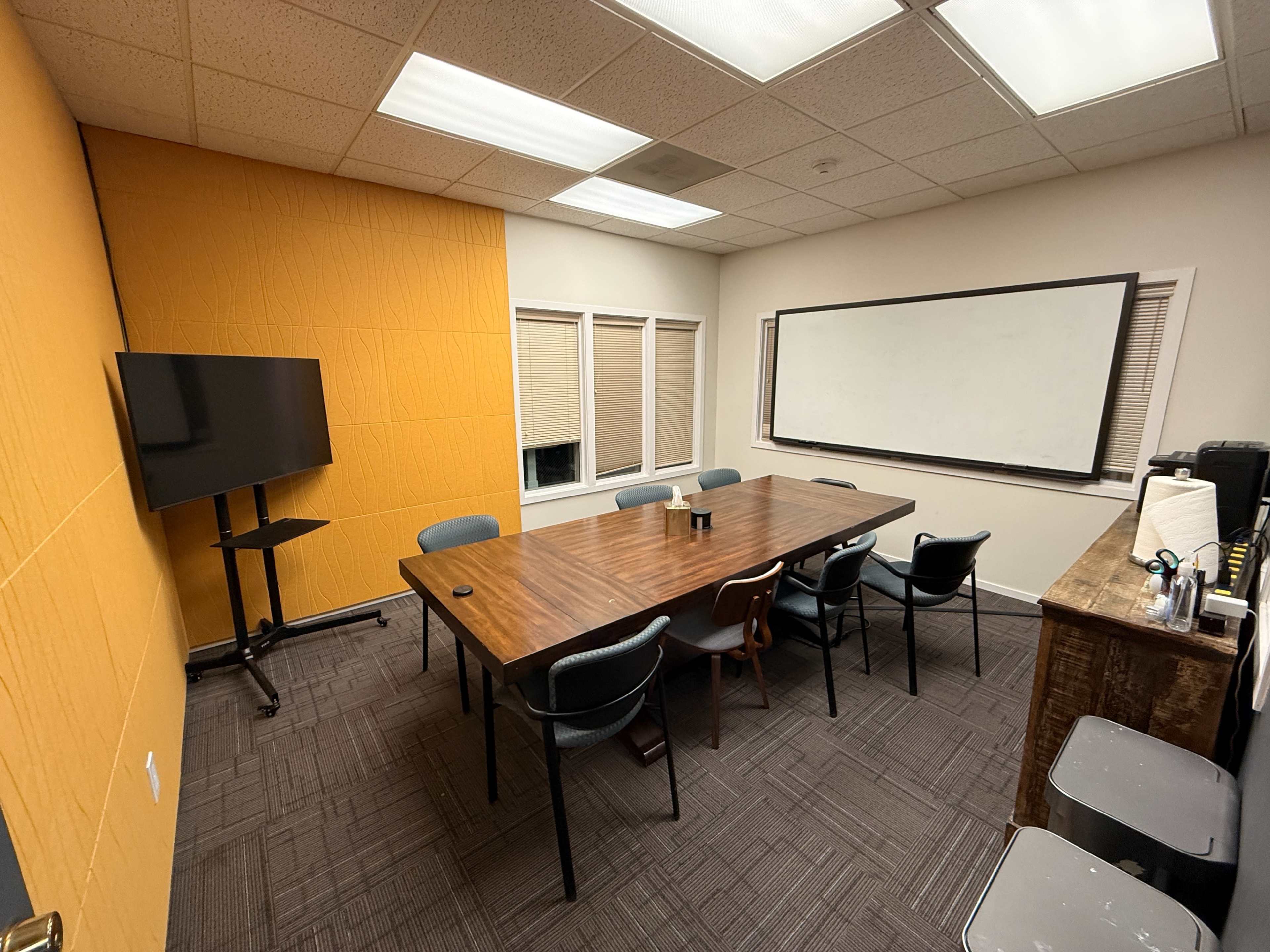 The image shows a conference room featuring a wooden table surrounded by chairs, a large screen on a stand, and a whiteboard on an adjacent wall.