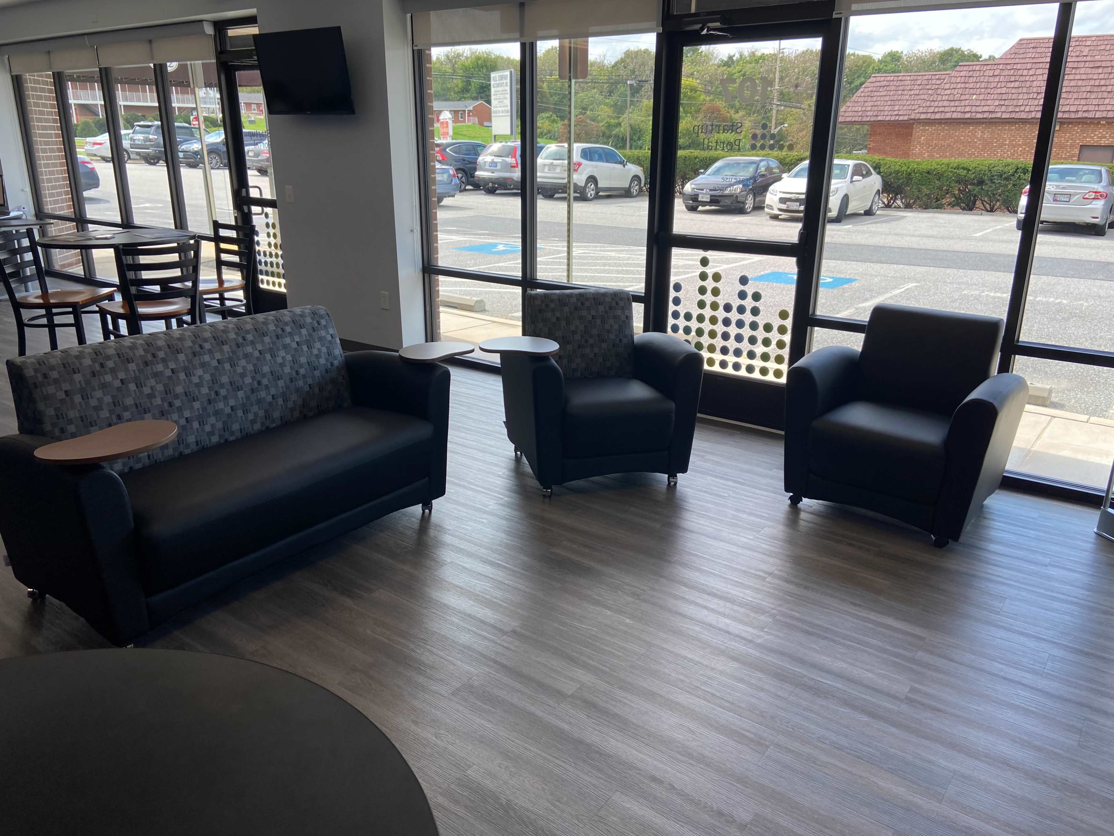 A seating area with two armchairs and a couch, all with small side tables, positioned near large windows overlooking a parking lot.
