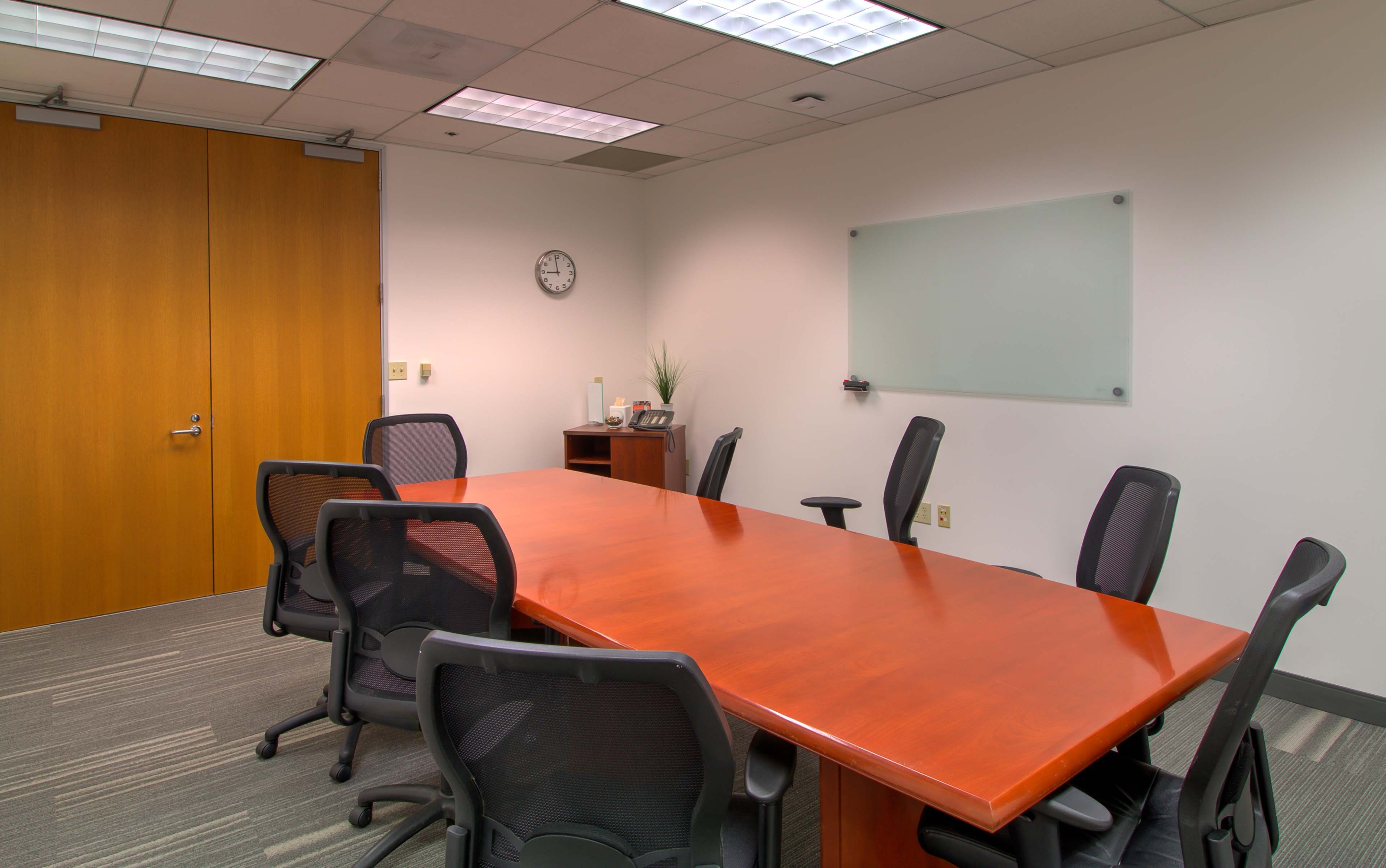 A large conference room features a wooden table surrounded by eight black chairs, with a wall clock and a whiteboard visible.