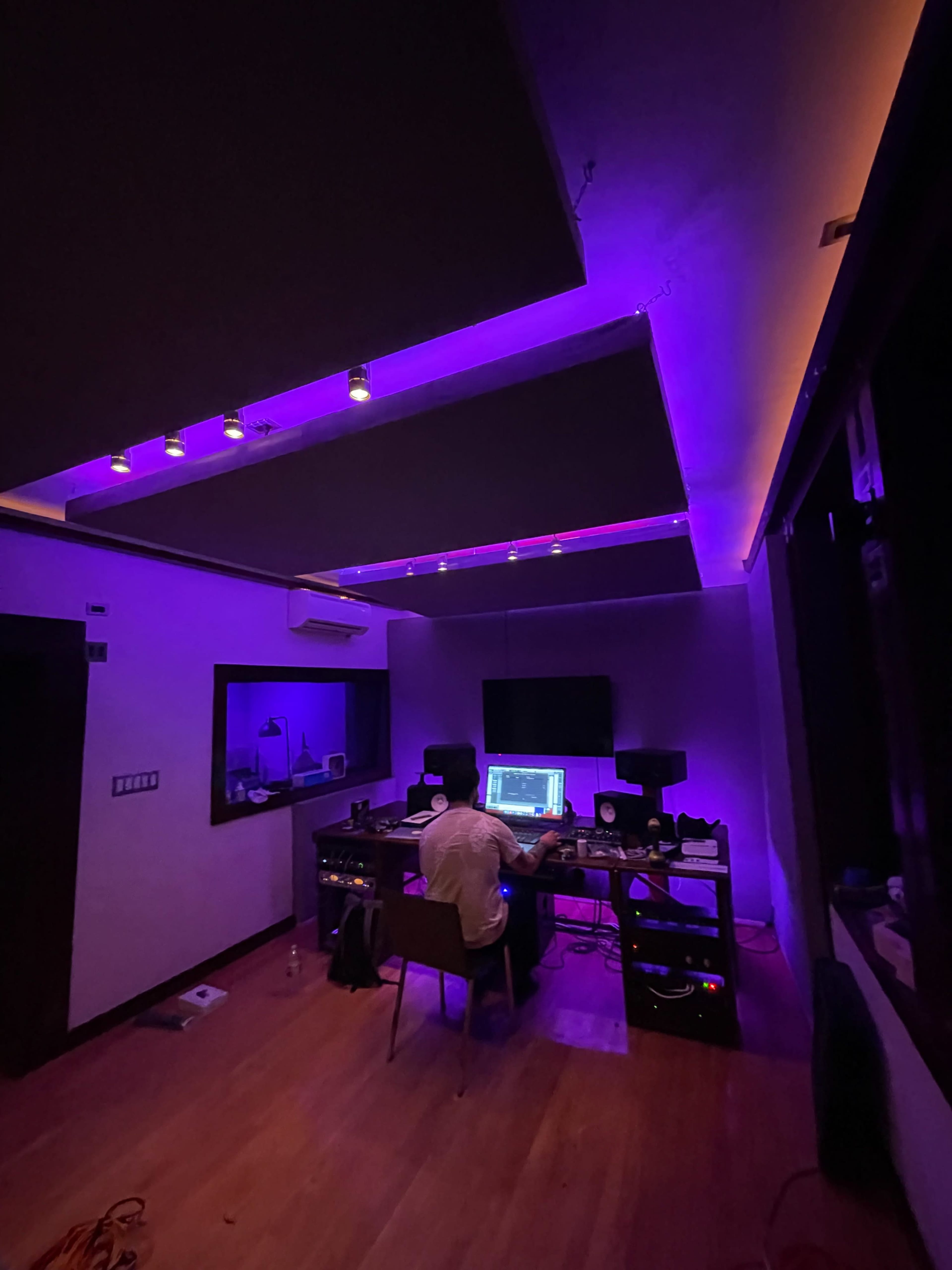 A person sits at a desk in a room illuminated by purple lights, surrounded by electronics and equipment.