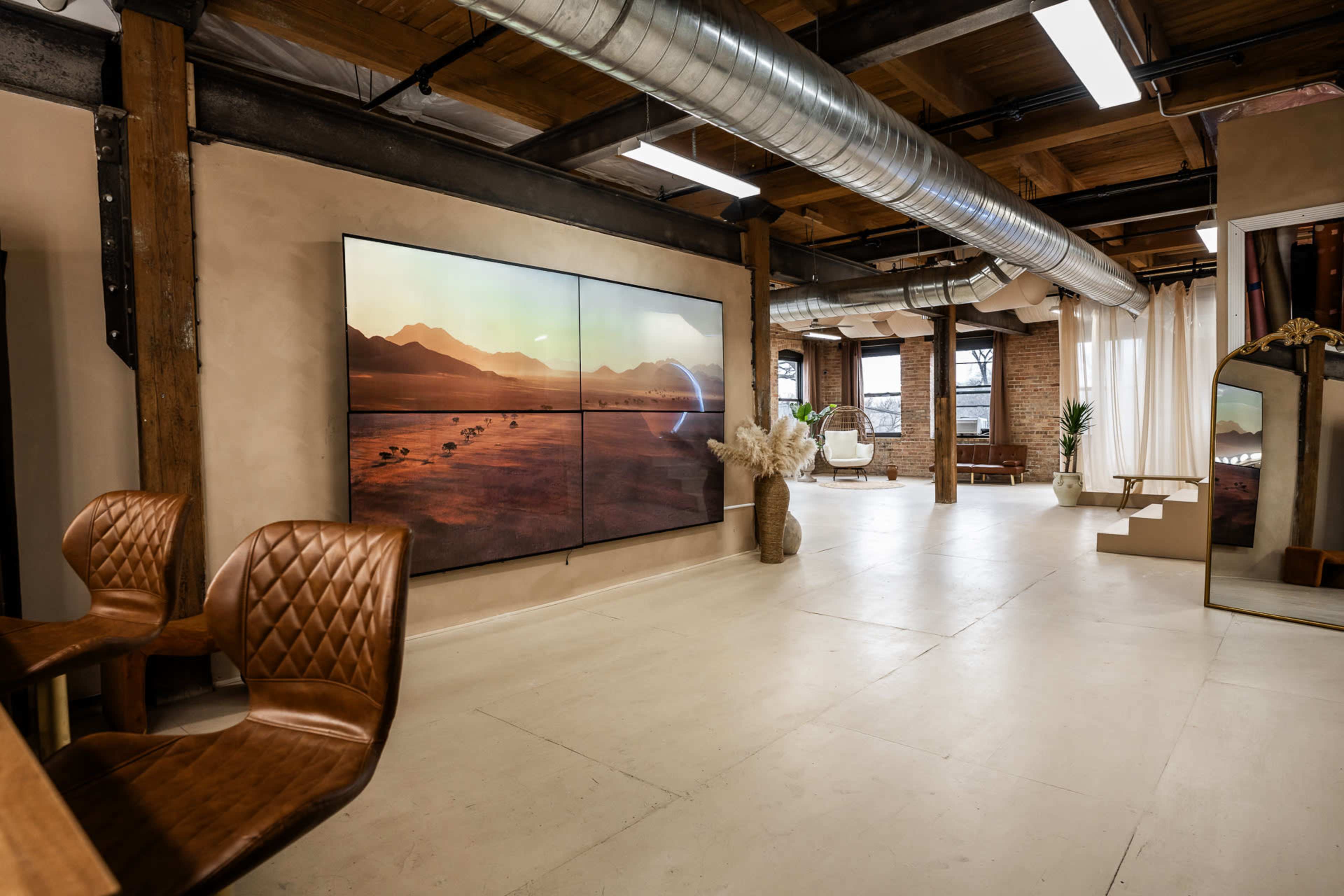 The image shows a modern interior space featuring a large, multi-panel screen displaying a desert landscape, with rustic wooden beams and industrial elements.