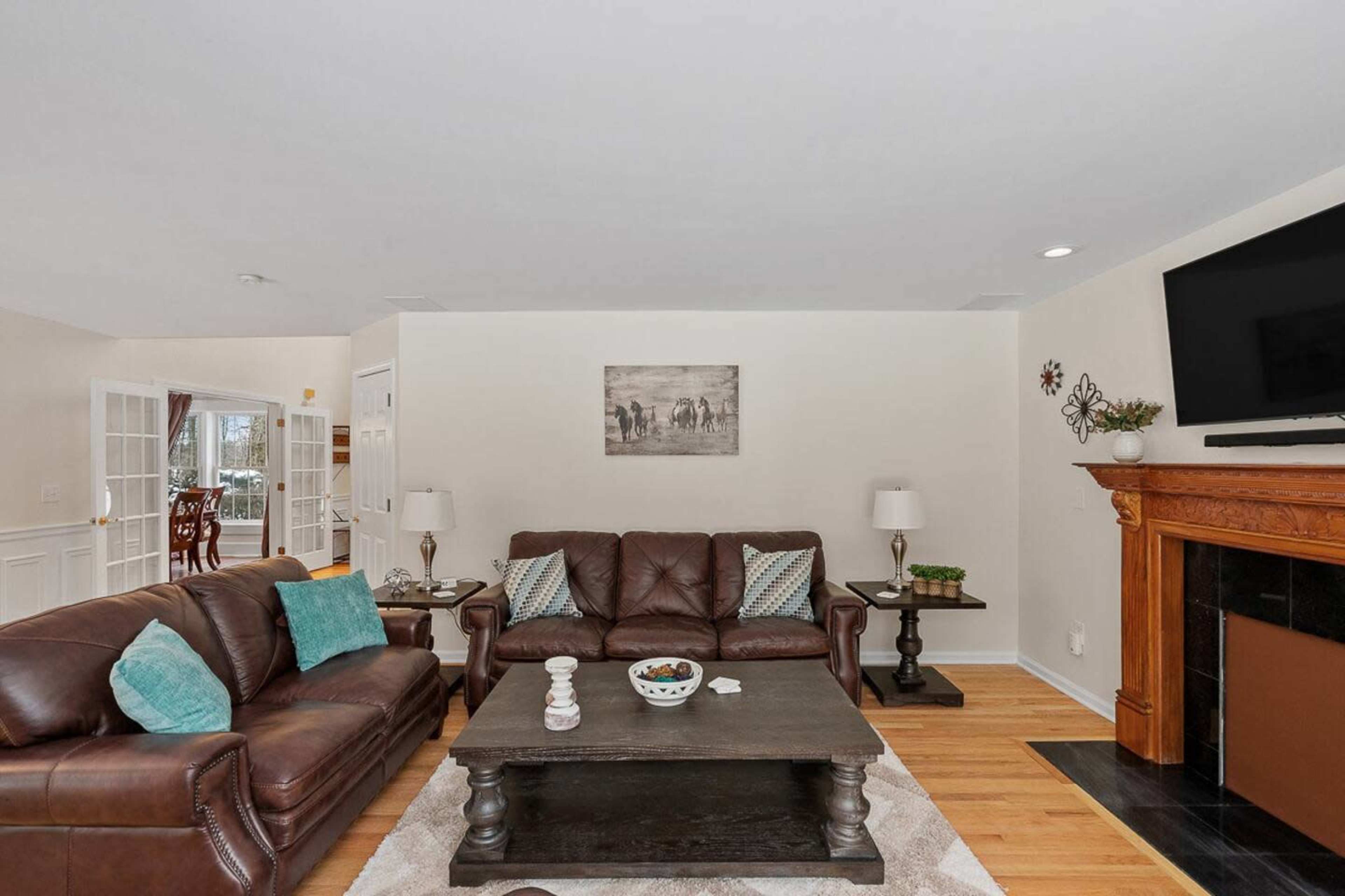 A living room features two brown leather sofas, a wooden coffee table, and a fireplace, with light-colored walls and a large flat-screen TV.