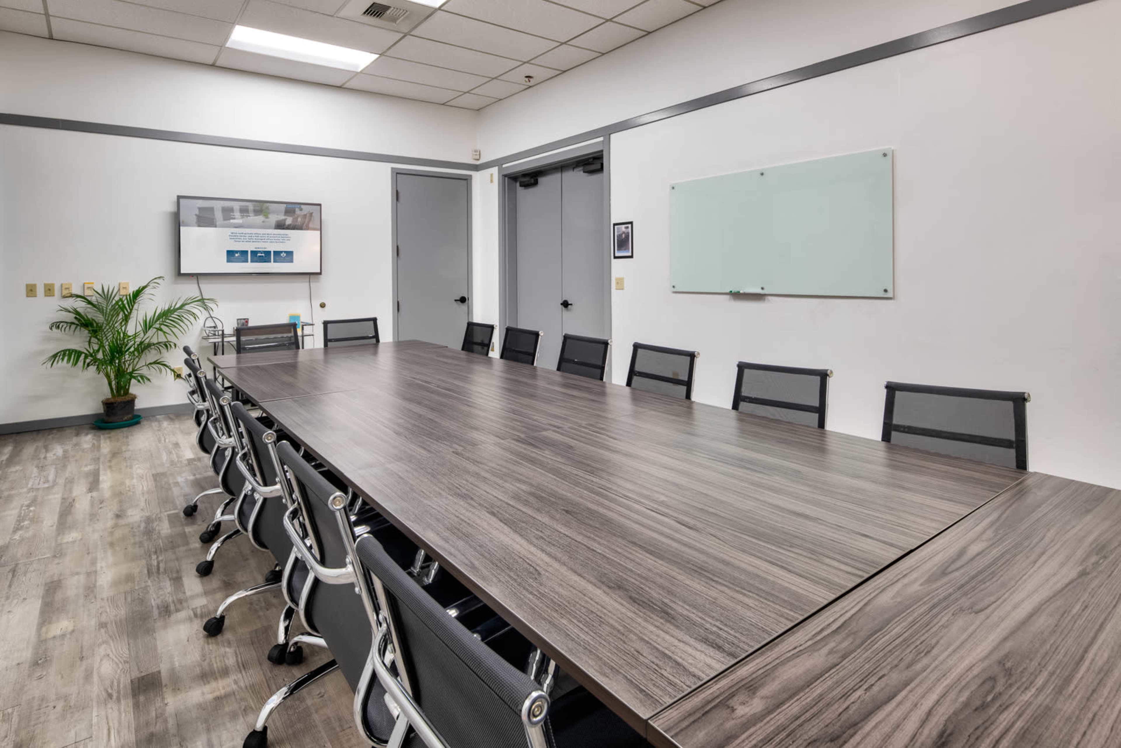 A conference room with a long wooden table surrounded by black mesh chairs, a whiteboard, and a wall-mounted screen.