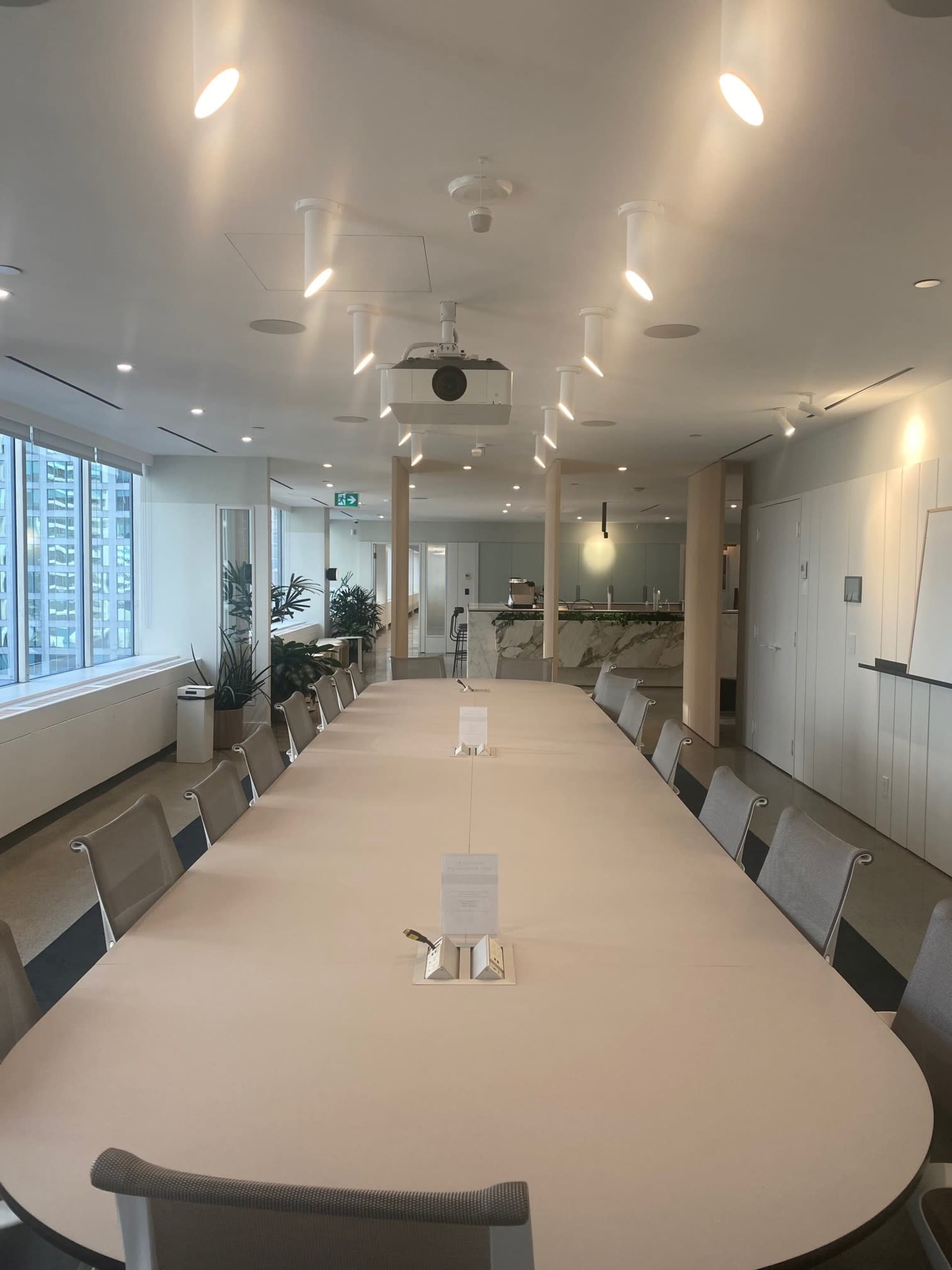 A large, modern conference room with a long oval table surrounded by chairs, windows allowing natural light, and a projector mounted on the ceiling.