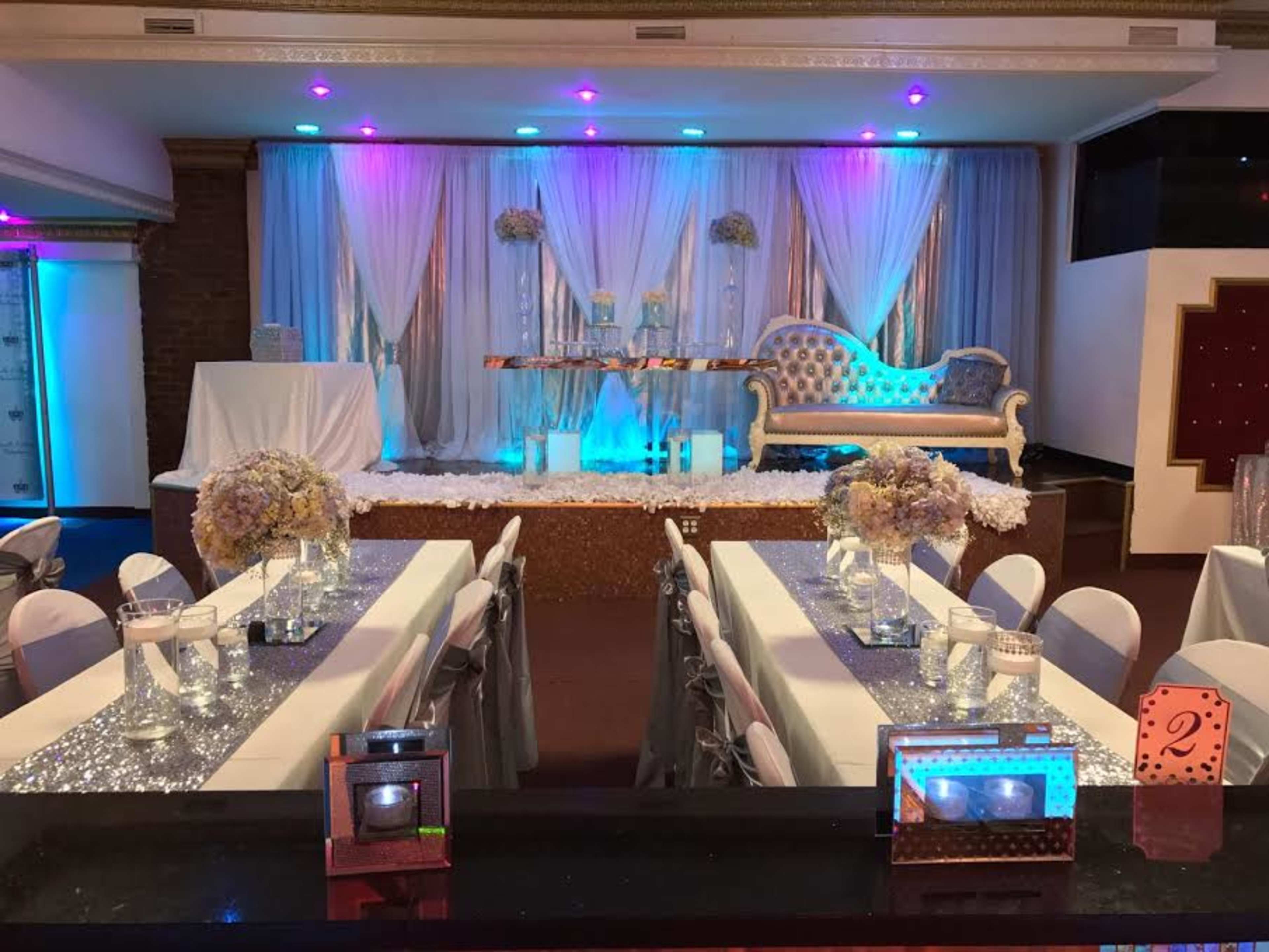 A banquet hall decorated with white and blue drapes, a stylized stage with floral arrangements, and tables set with elegant centerpieces.