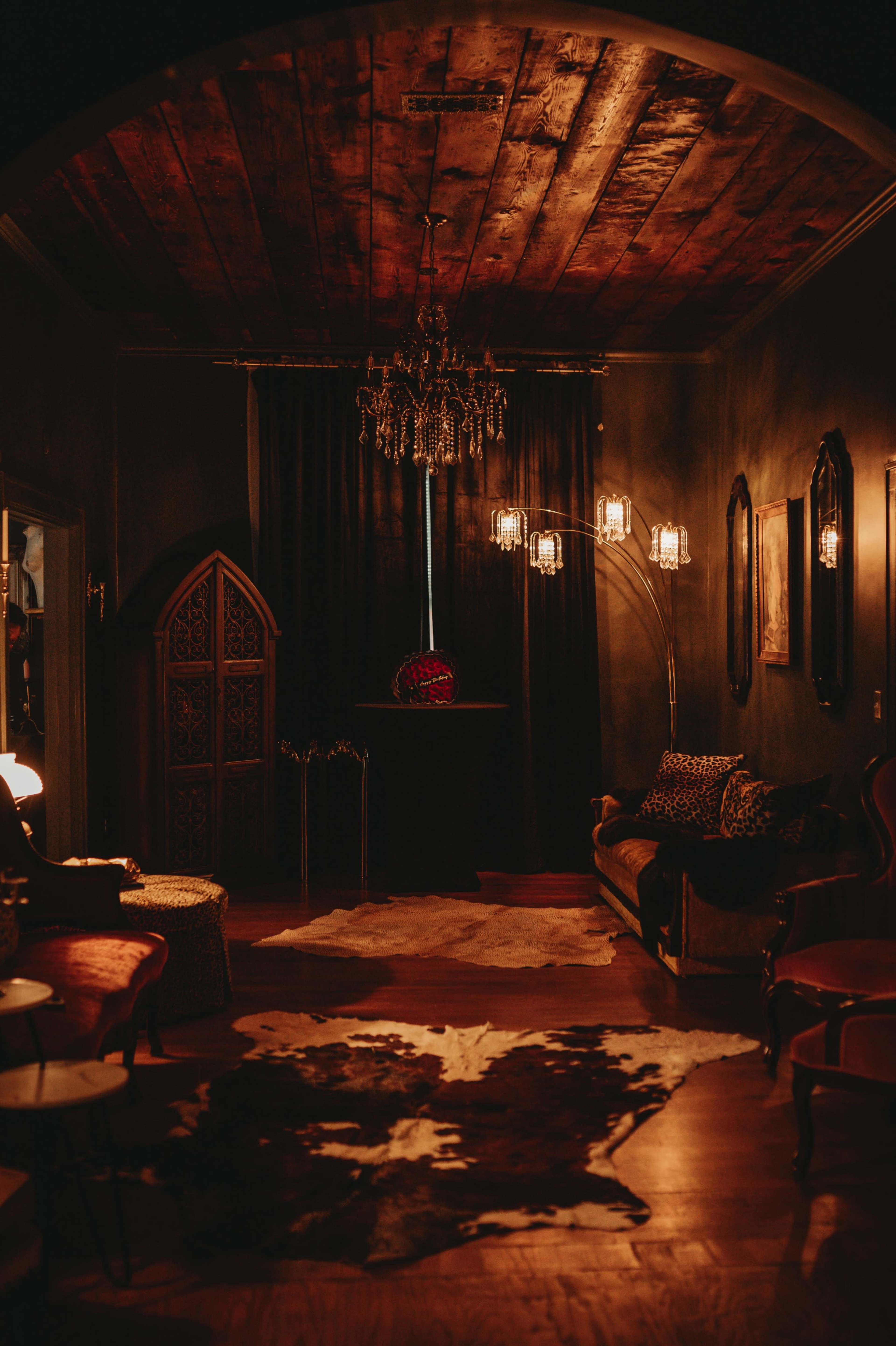 The image shows a dimly lit room with a wooden ceiling, elegant furniture, a cowhide rug on the floor, and decorative lighting fixtures.