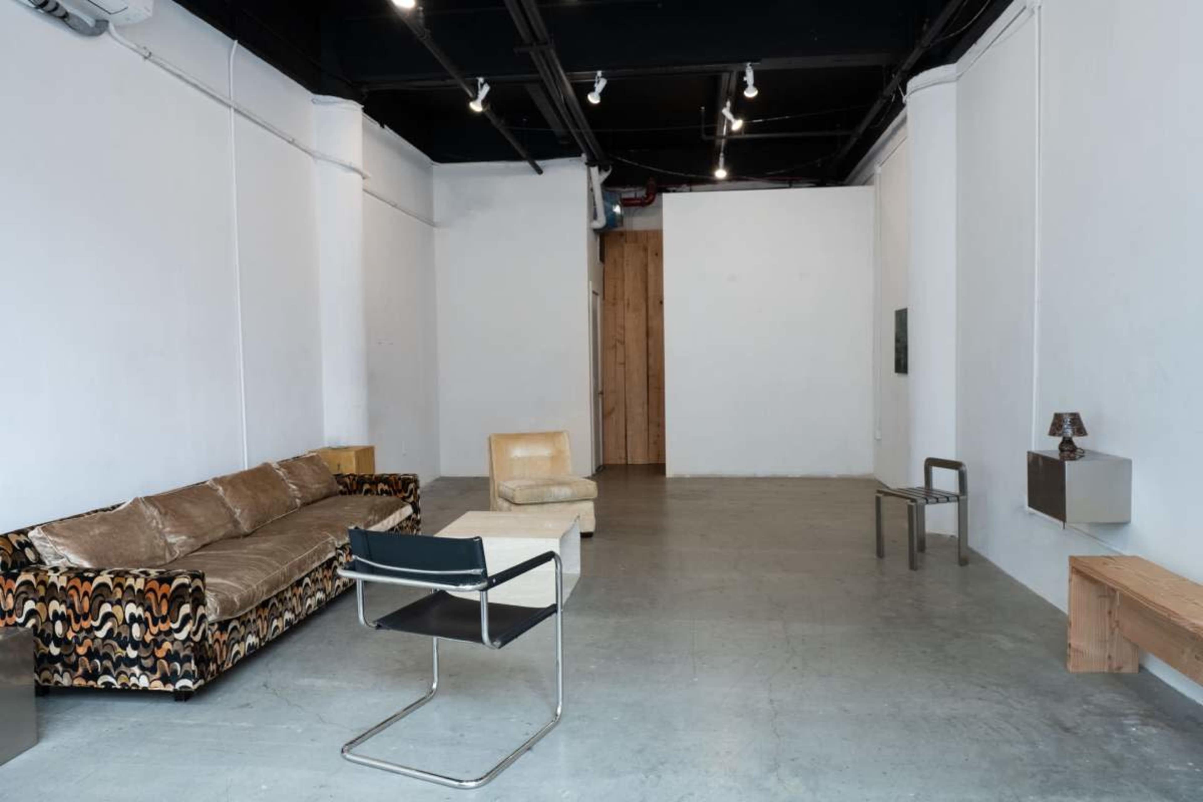 The image shows a minimalist interior space featuring a patterned sofa, a simple chair, a coffee table, and wooden accents along the walls.
