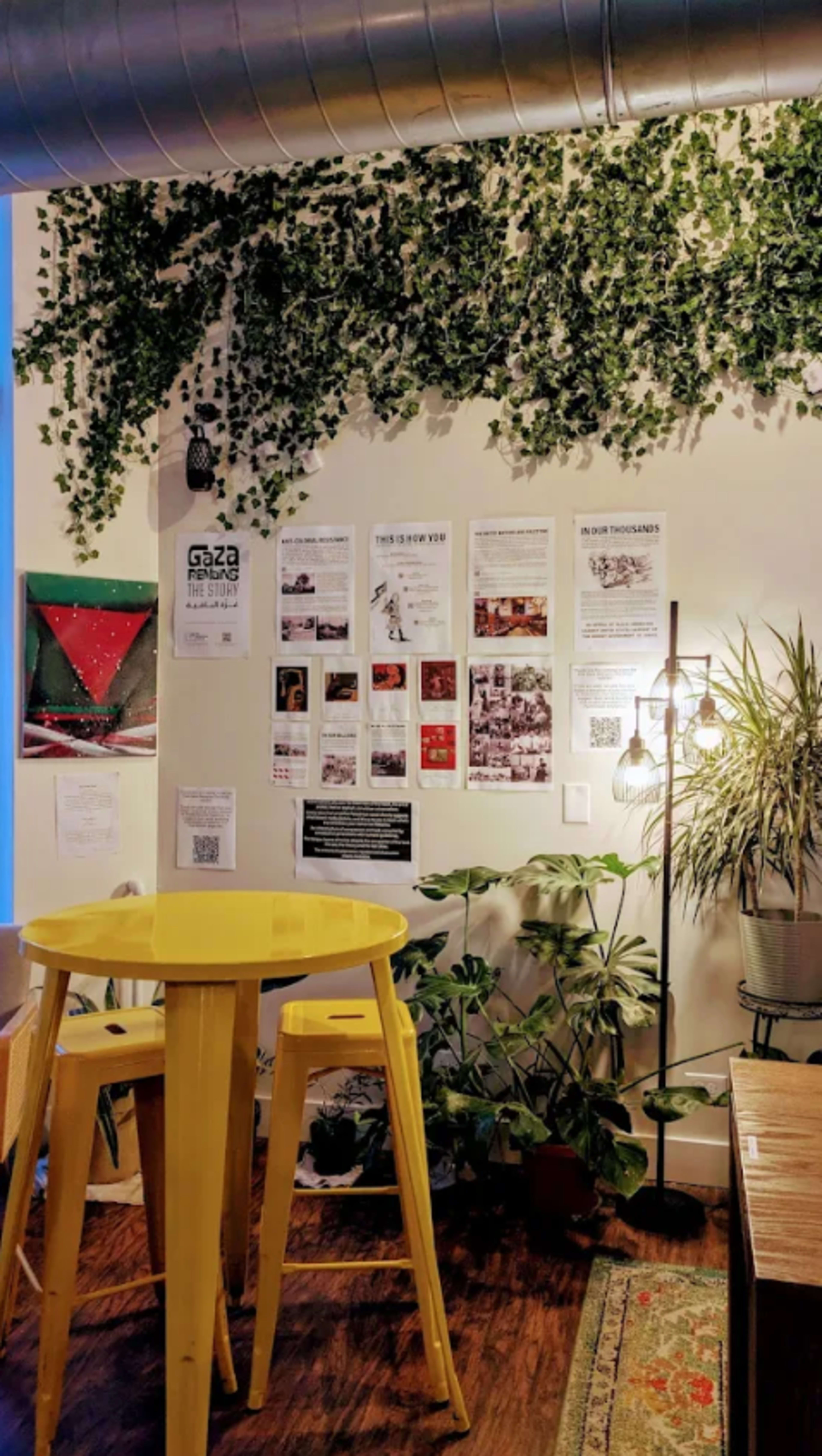 A yellow round table with two stools stands in front of a wall covered in green leaves and various posters.