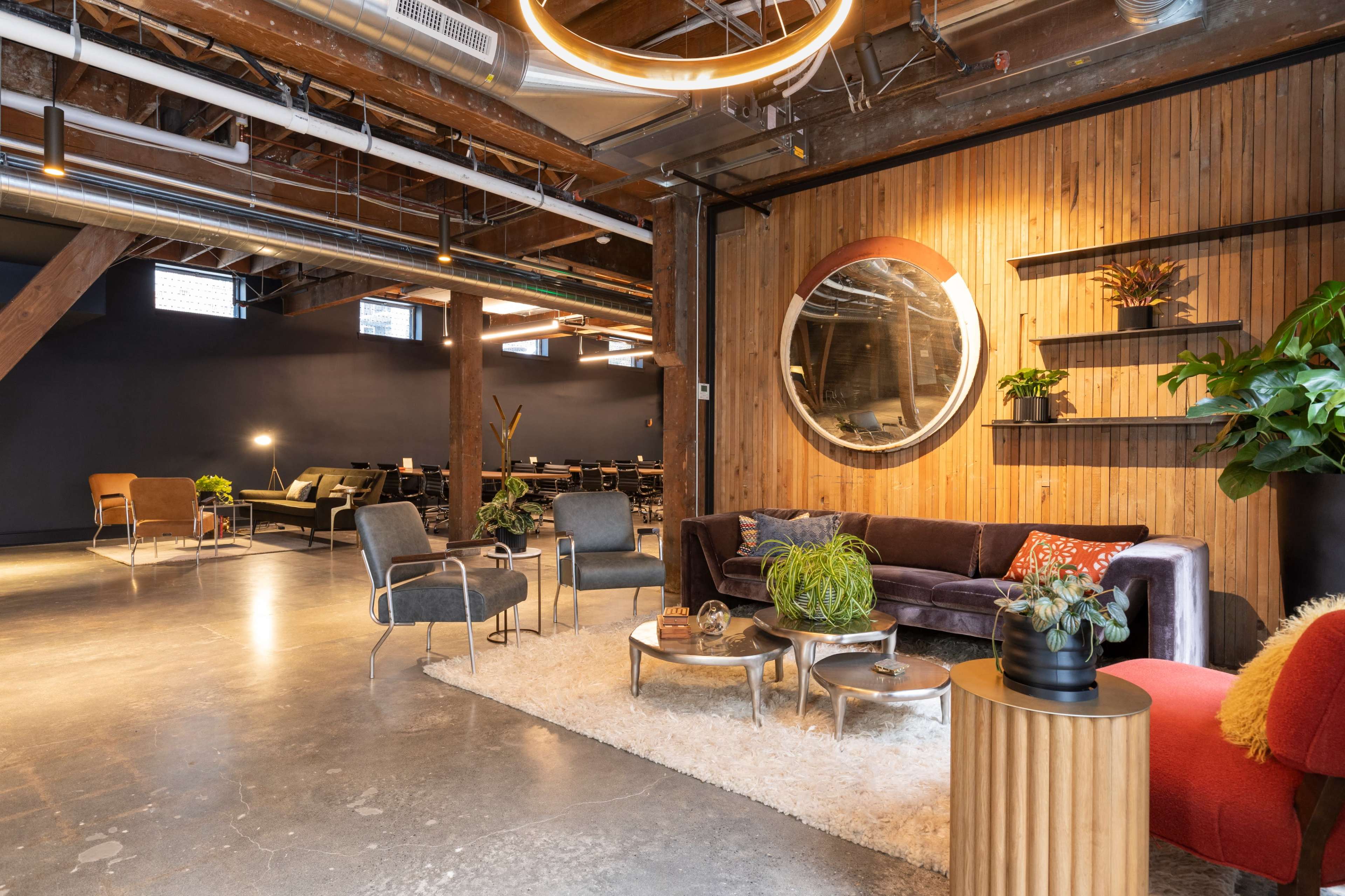A modern open workspace with a cozy seating area featuring a velvet couch, chairs, and various plants, complemented by a wooden accent wall and industrial-style lighting.