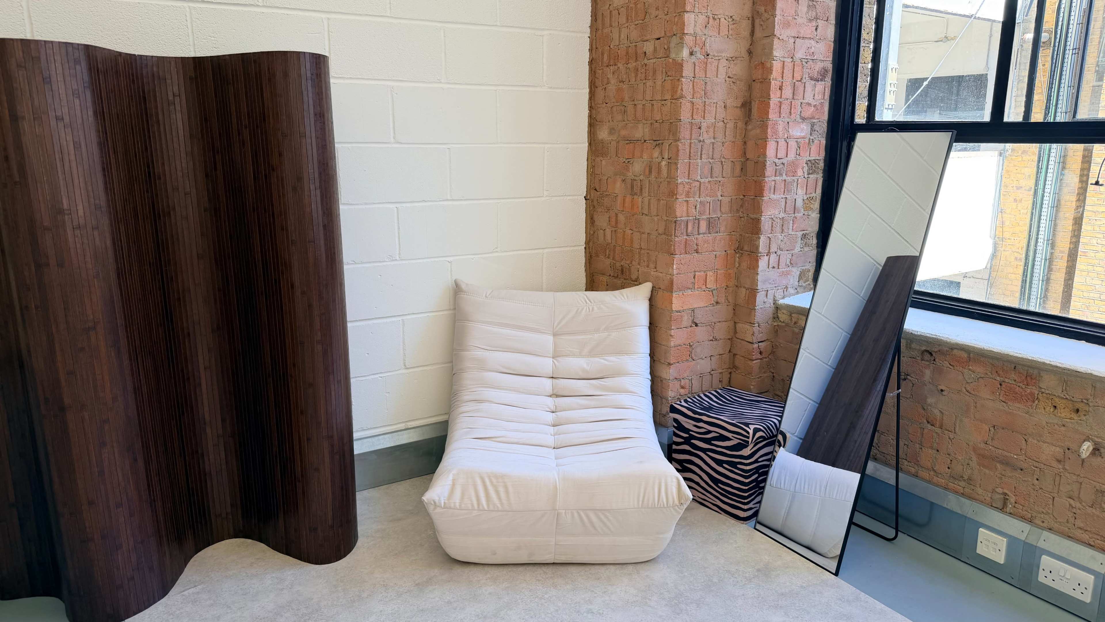 The scene features a casual seating cushion, a large floor mirror, and a decorative divider against a textured brick wall.
