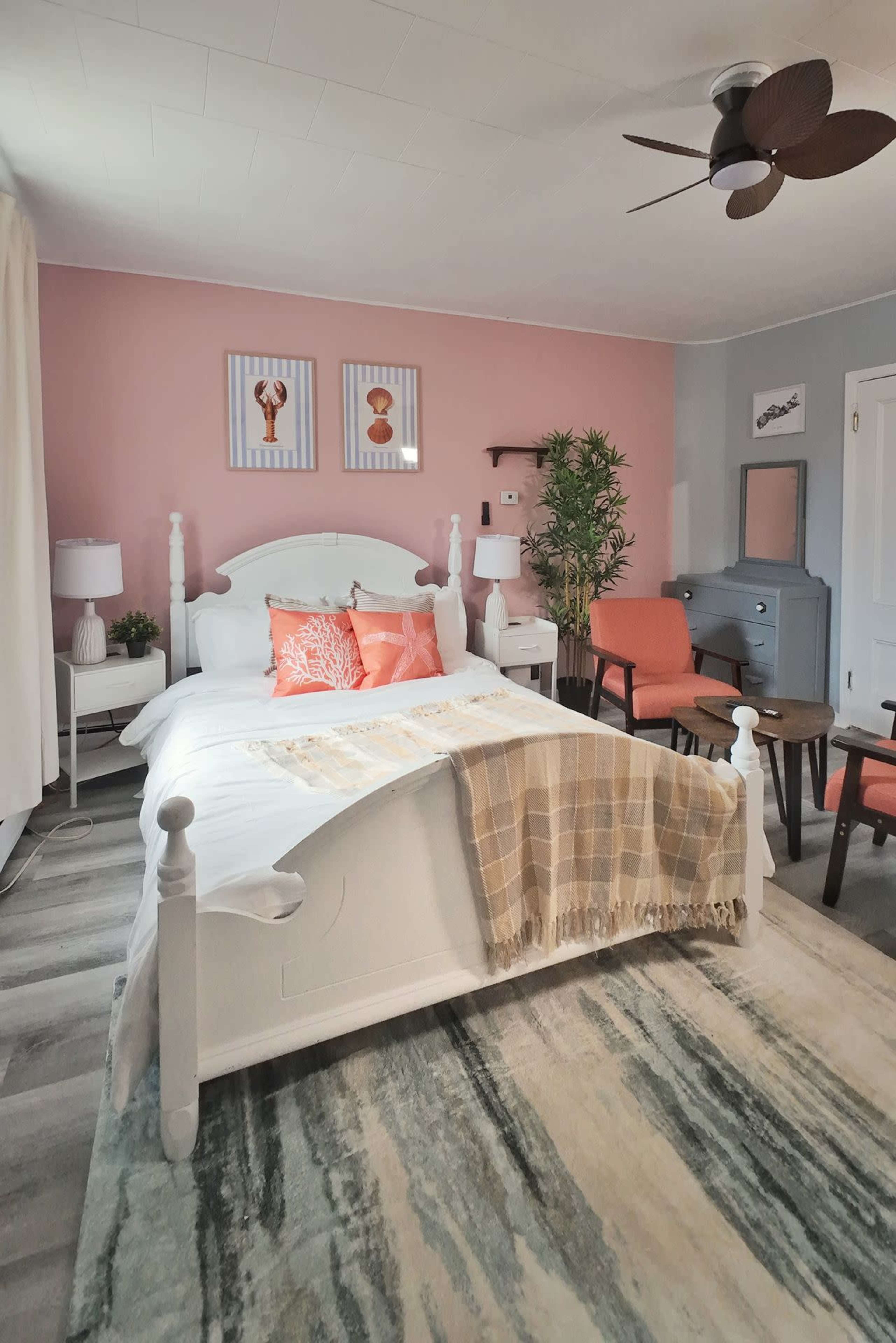 The room features a white bed with decorative pillows, two orange chairs, a small wooden table, and artwork on the wall, all set against a pink accent wall.
