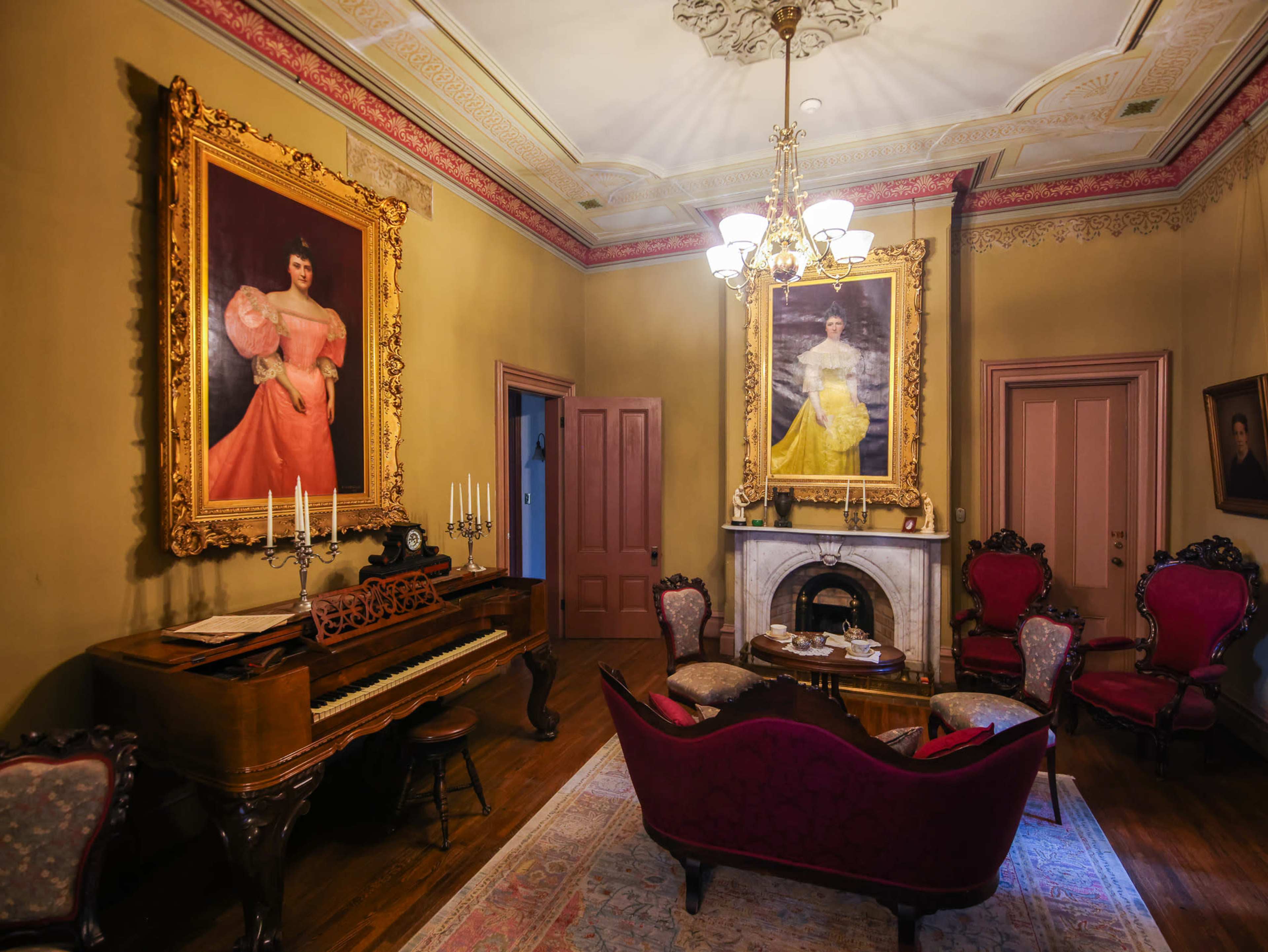 The room features a grand piano, ornate framed portraits on the walls, and vintage furniture arranged around a fireplace.