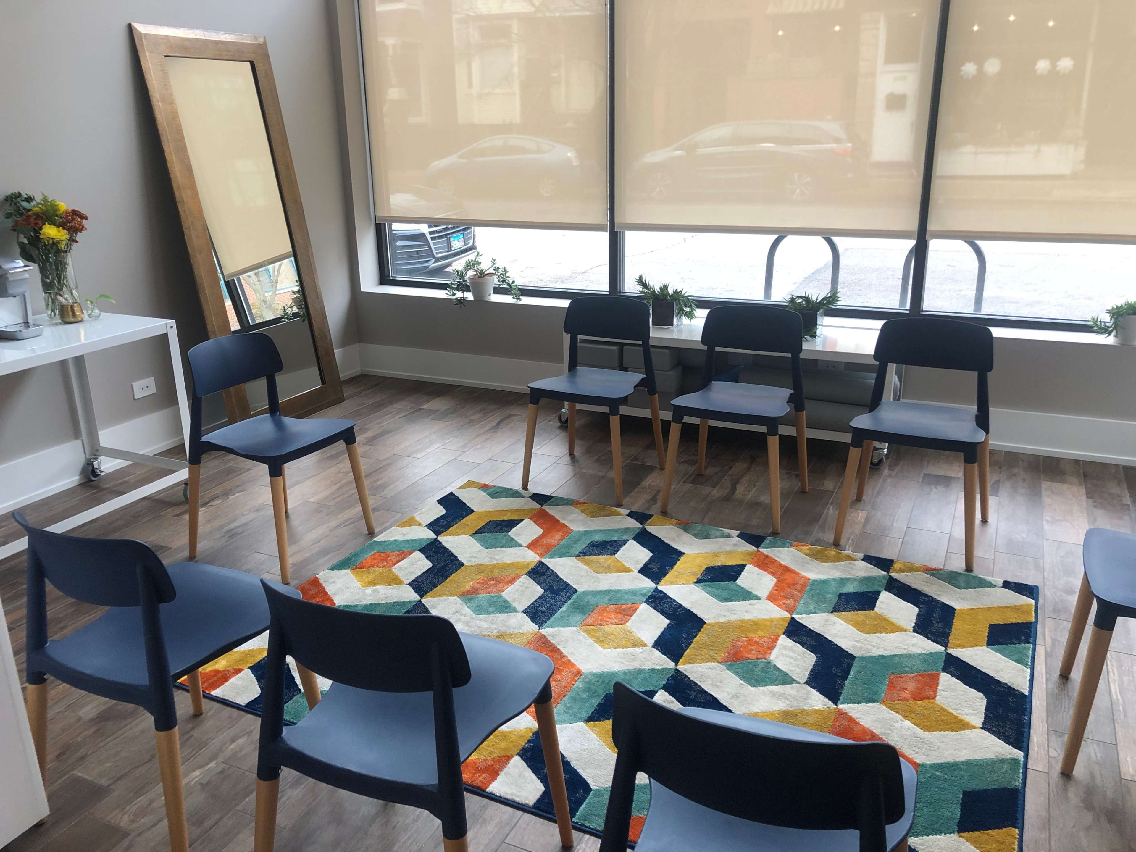 A circle of blue chairs surrounds a patterned rug in a brightly lit room with large windows and a mirror.