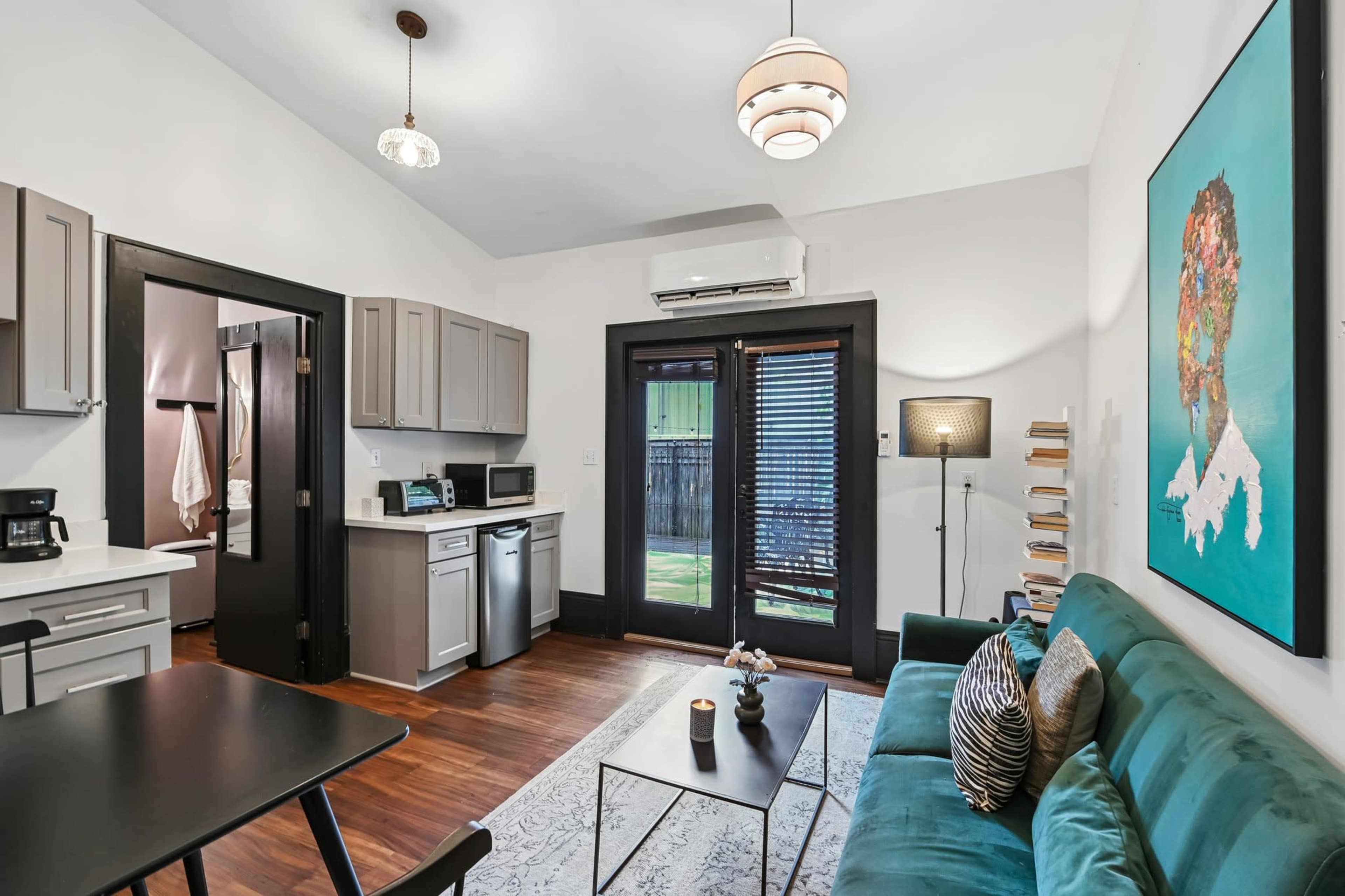 The image shows a modern living area with a green sofa, a dining table, and an open kitchen featuring gray cabinets and a coffee maker, all illuminated by natural light through sliding glass doors.