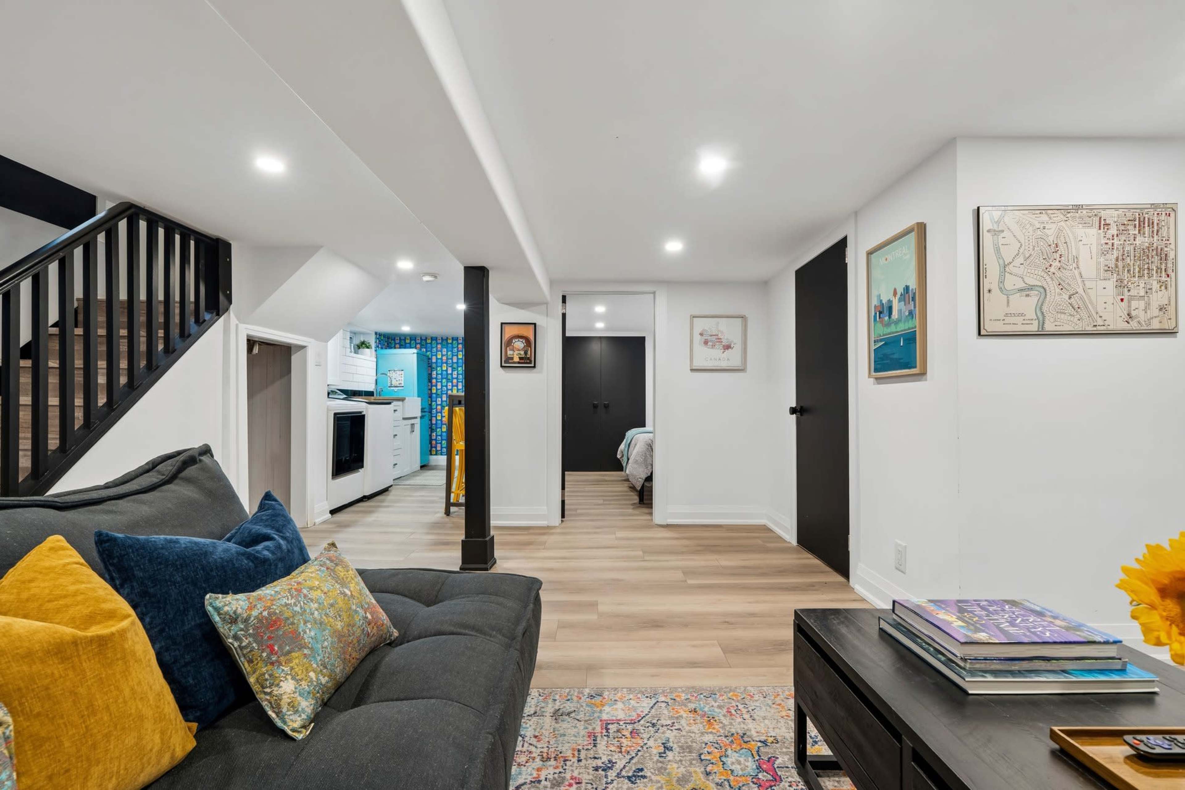 A well-lit basement living area features a gray couch with colorful pillows, wooden flooring, and a glimpse of a kitchen and bedroom in the background.